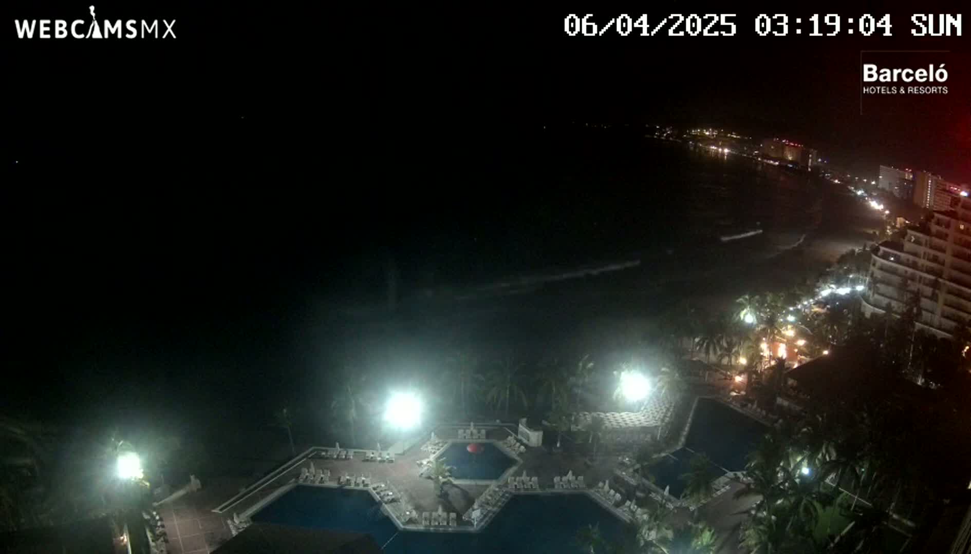 A nighttime, overhead view of a resort pool area with palm trees and a brightly lit coastline in the distance.