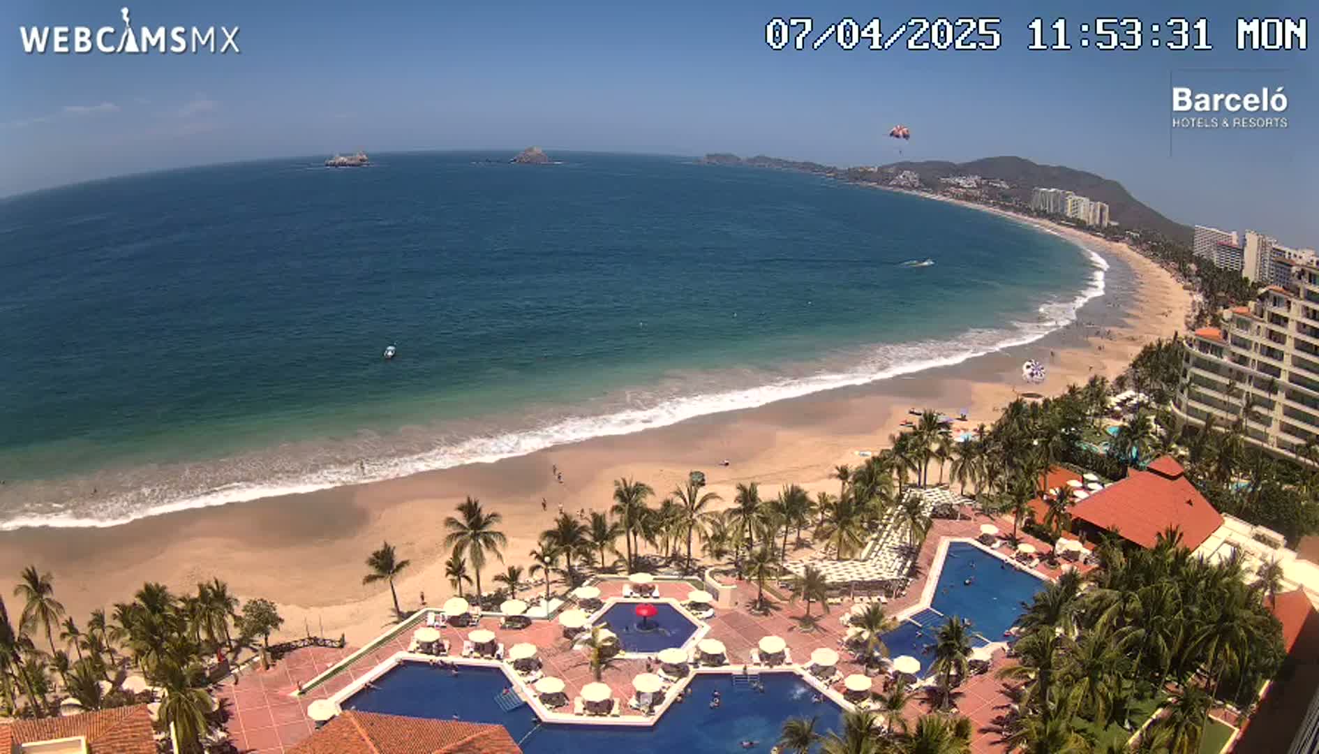 A beach resort with multiple pools is situated on a sandy beach next to a calm, blue ocean under a sunny sky.