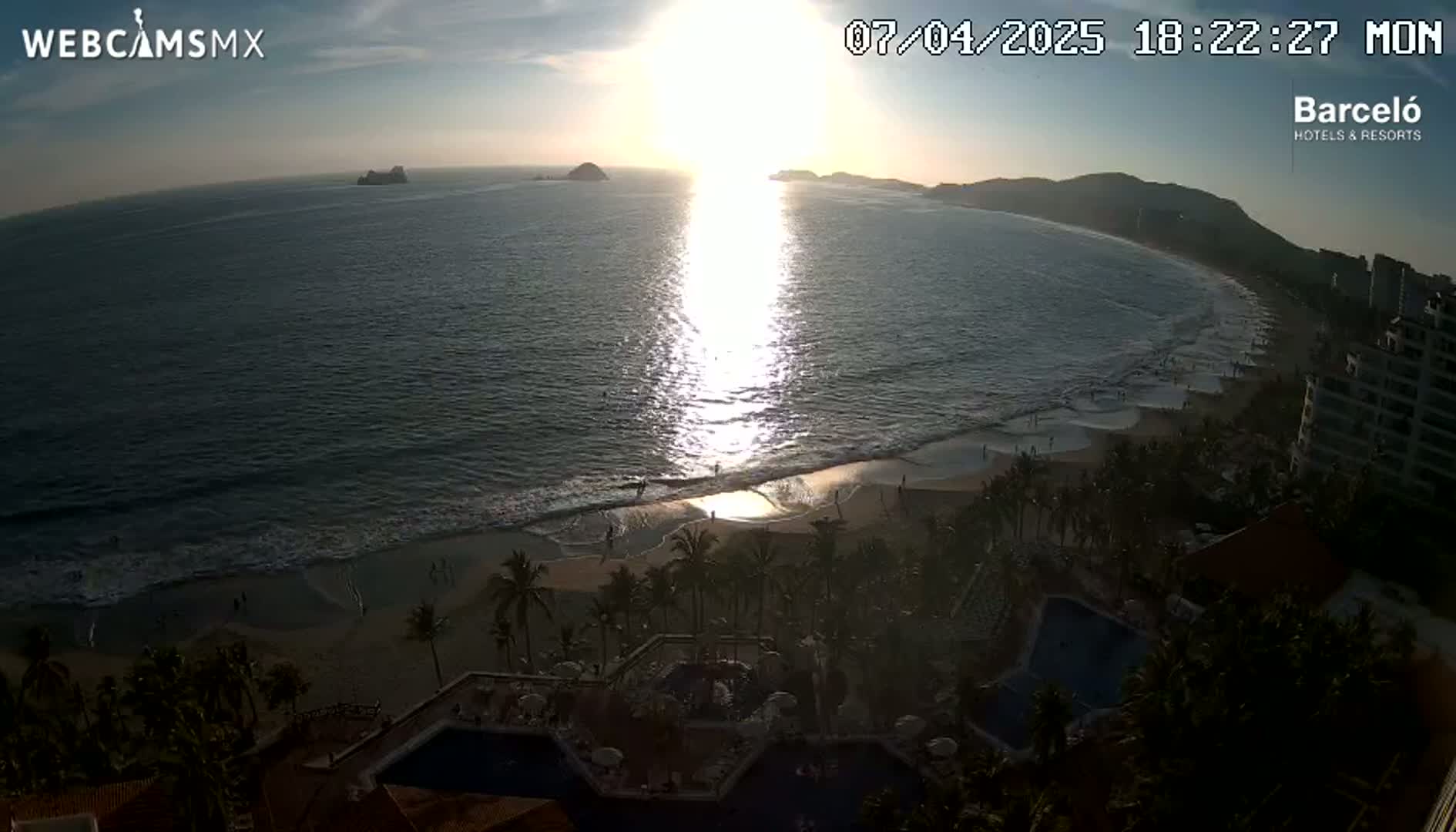 An ocean beach at sunset, with palm trees and buildings visible along the shore, and a calm sea reflecting the sun's light.