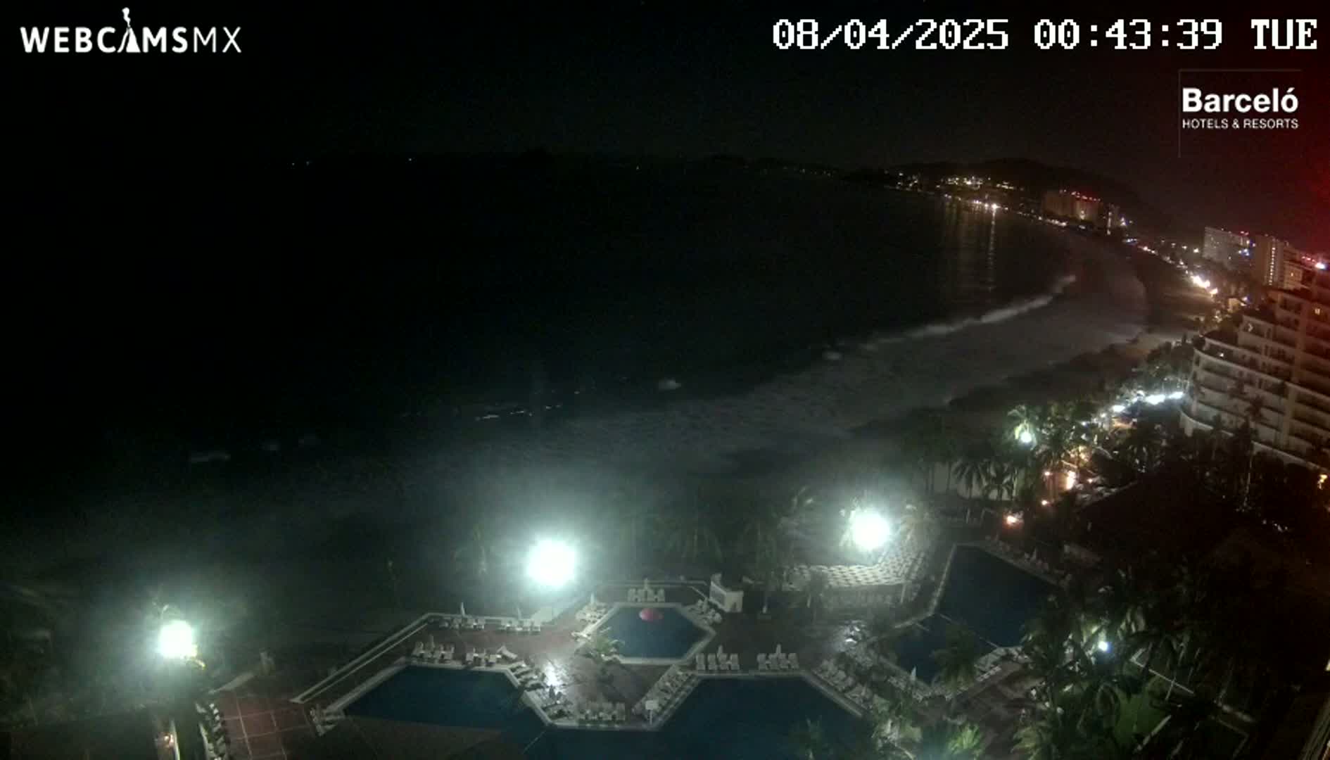 A nighttime view of a resort complex with illuminated pools overlooking a dark ocean with waves under a clear sky.