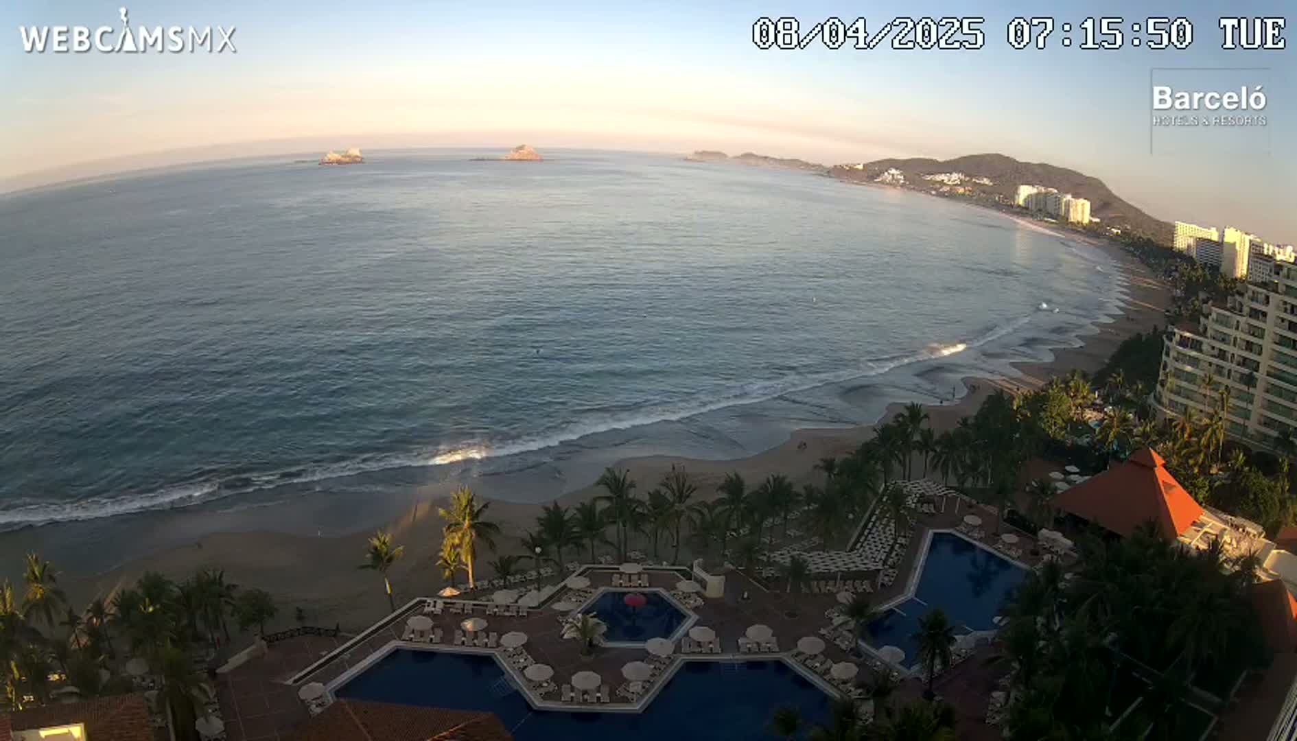 An aerial view of a resort with multiple pools overlooking a beach and ocean under a clear, sunrise sky.