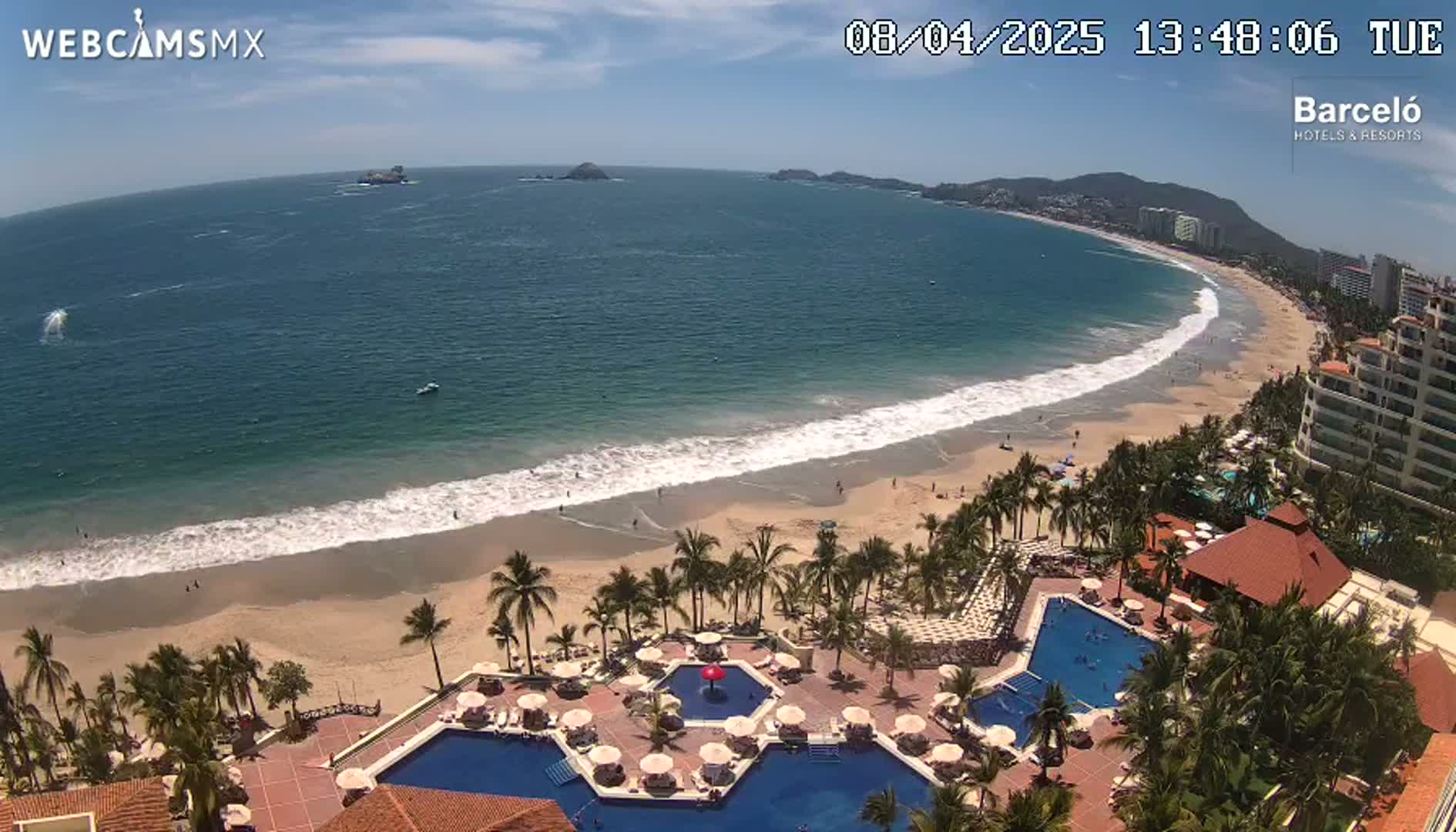 An aerial view shows a beach with light-colored sand and gentle waves lapping the shore under a sunny sky,  with a resort featuring multiple pools and palm trees situated on the beachfront.