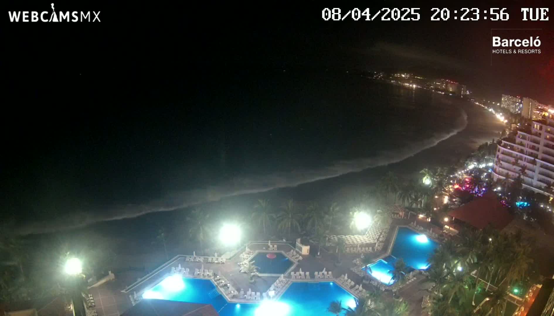 An illuminated resort pool area overlooks a dark ocean beach with gentle waves under a nighttime sky.