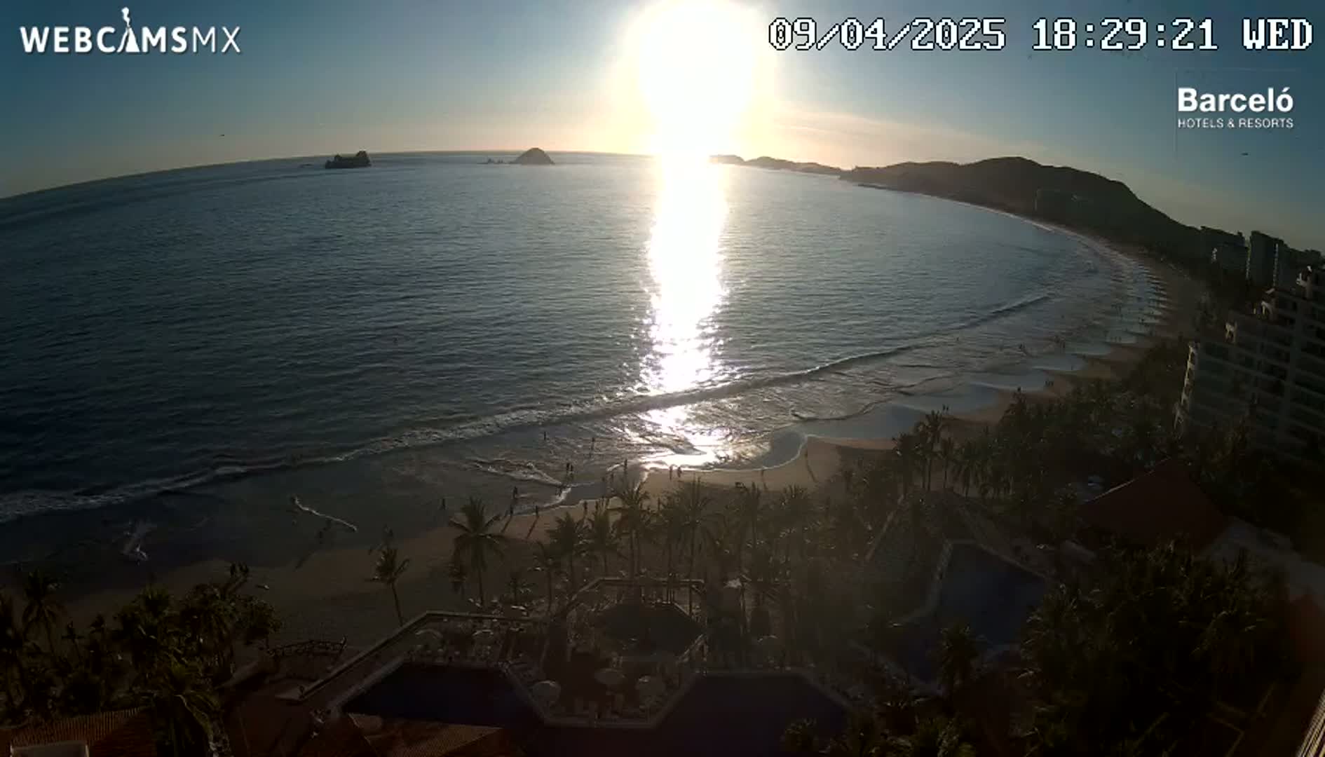 A beach scene at sunset shows the sun reflecting on the ocean, waves lapping the shore, palm trees lining the sand, and buildings in the background.
