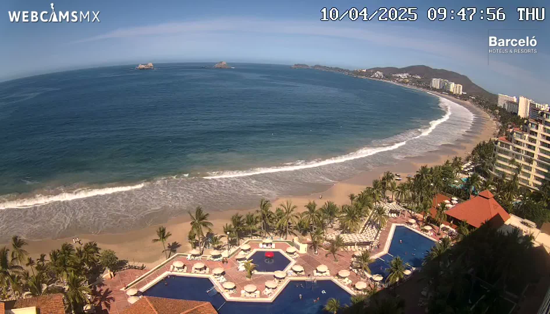 An aerial view shows a resort with multiple pools overlooking a sandy beach and ocean with gentle waves under a clear, sunny sky.