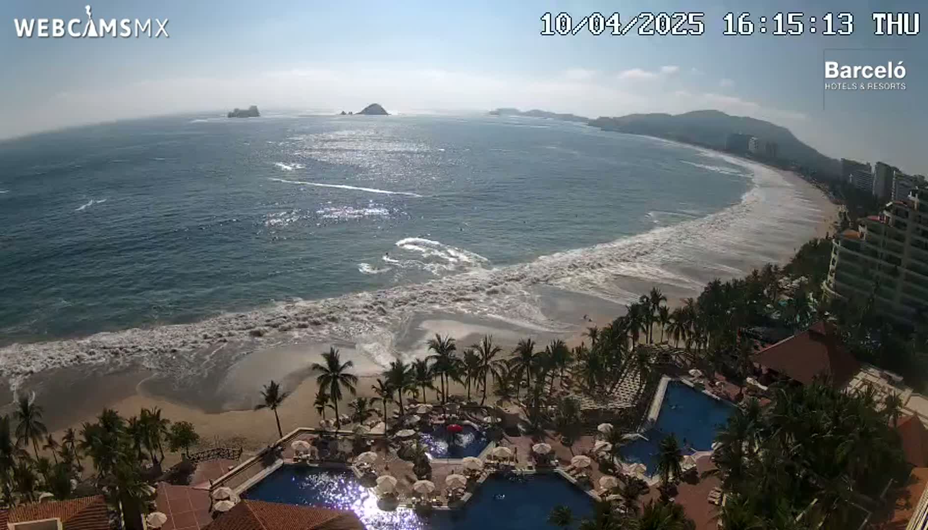 An ocean beach with moderate waves is seen from an elevated vantage point overlooking a resort with multiple swimming pools surrounded by palm trees under a mostly sunny sky.