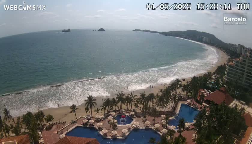 An aerial view shows a resort with multiple pools overlooking a sandy beach with waves rolling in under a mostly sunny sky.