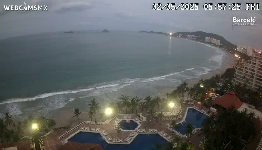 An overcast, early morning view shows a resort with multiple pools overlooking a sandy beach and calm ocean waves.