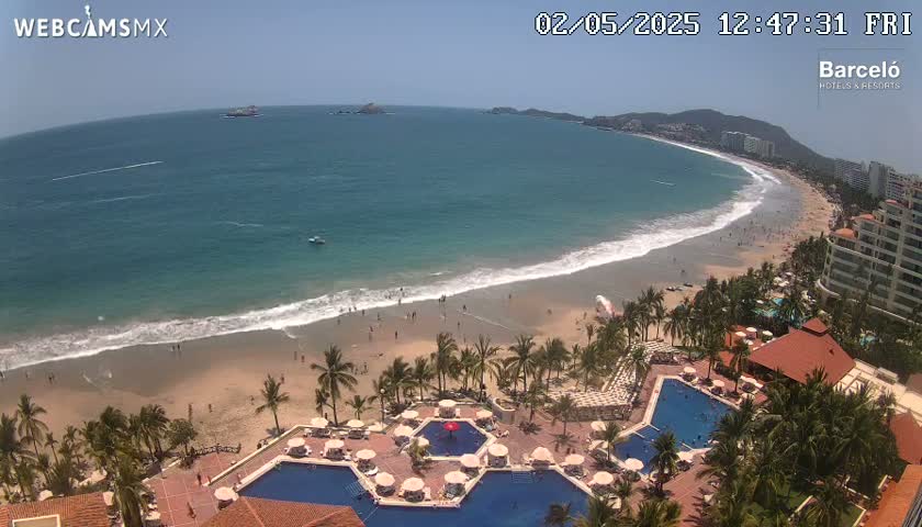 An aerial view shows a beach with a resort featuring multiple pools and palm trees under sunny skies, with calm waves lapping the shore.
