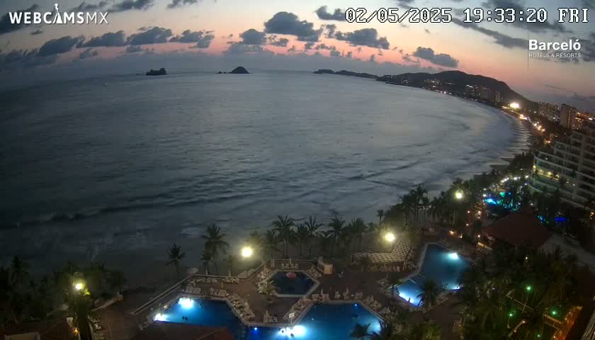 An oceanfront resort at twilight, with calm waves lapping the shore and multiple illuminated pools surrounded by palm trees.
