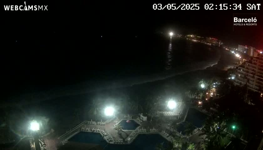 A nighttime view of a resort complex with multiple pools, illuminated by artificial lights, overlooking a dark ocean under a clear sky.