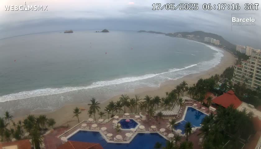 An aerial view shows a beach with gentle waves under an overcast sky, overlooking a resort with multiple pools and palm trees.