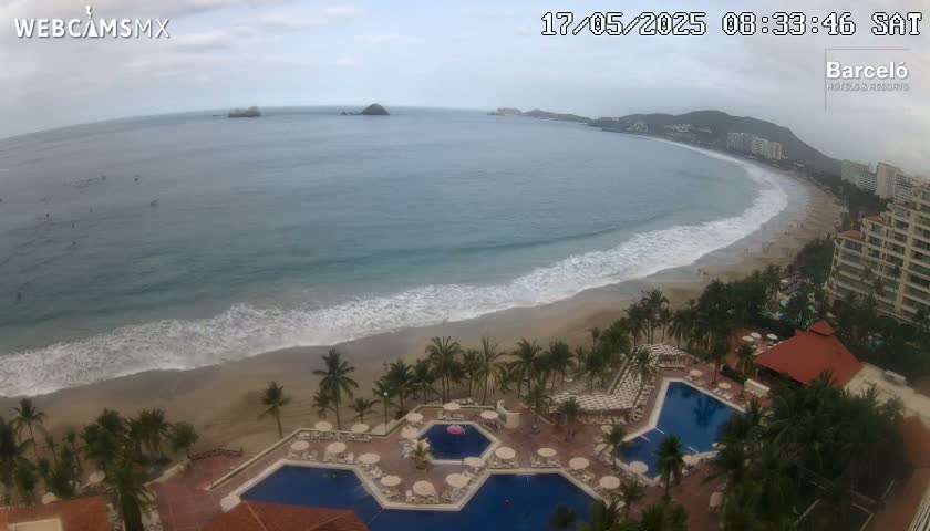 An aerial view shows a resort with multiple swimming pools situated on a sandy beach next to a calm ocean with small waves under an overcast sky.