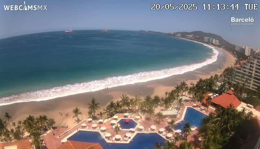An aerial view shows a resort with multiple pools overlooking a sandy beach and ocean with gentle waves under a clear, sunny sky.