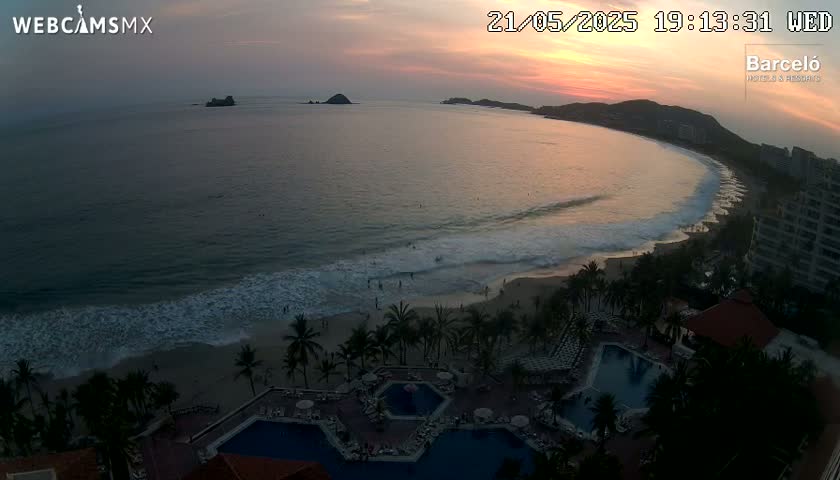 A sunset over a crescent-shaped beach with gentle waves, palm trees, and resort pools.