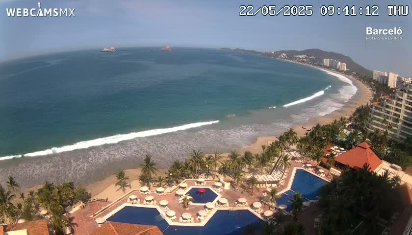 An aerial view shows a resort with multiple pools overlooking a sandy beach and ocean with gentle waves under a mostly sunny sky.