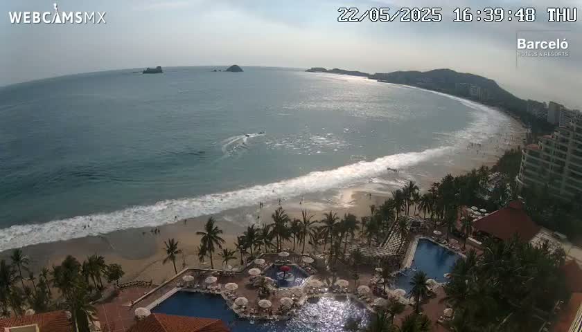 An aerial view shows a beach with gentle waves under a partly cloudy sky, overlooking a resort with multiple pools and palm trees.
