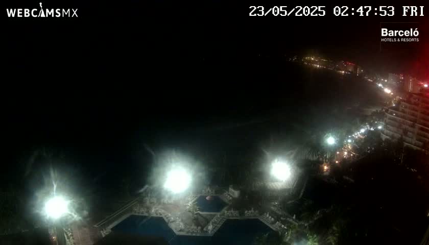 A nighttime, high-angle view shows a resort with several illuminated pools and surrounding buildings, extending to a dimly lit beachfront and city lights beyond.