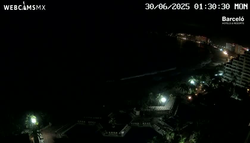 A nighttime, high-angle view shows a resort pool area and a dark ocean with a dimly lit coastline in the distance.