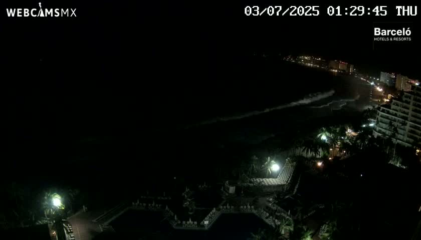 A nighttime view of a resort area near a beach with dark, wavy ocean and some city lights visible in the distance.