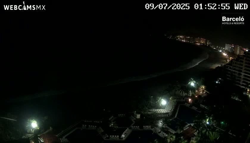 A nighttime, overhead view of a resort area showing pools, palm trees, and a dark ocean beach with dimly lit buildings in the distance.