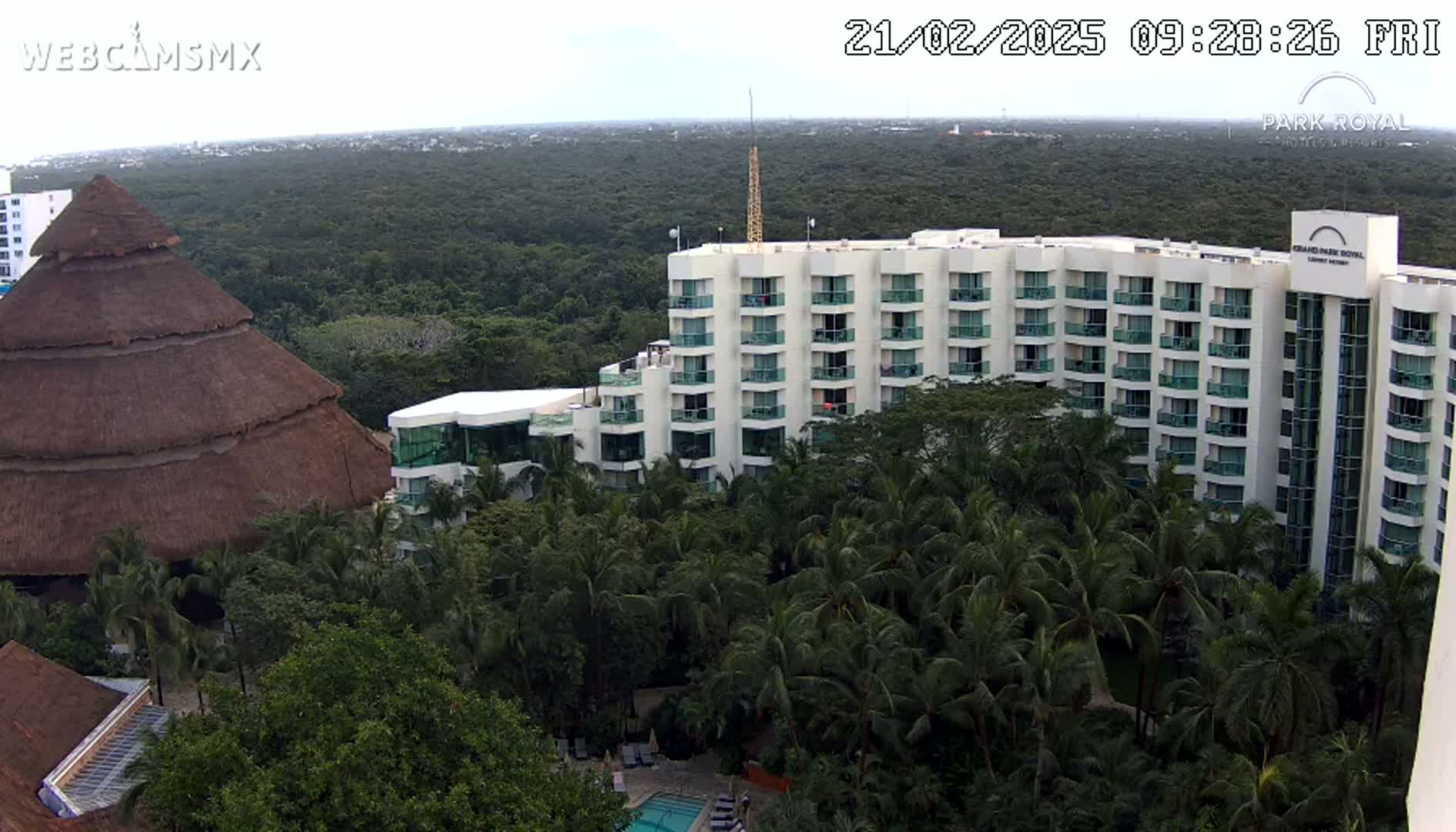 A large, multi-story hotel building is nestled among lush green tropical vegetation under a mostly cloudy sky.