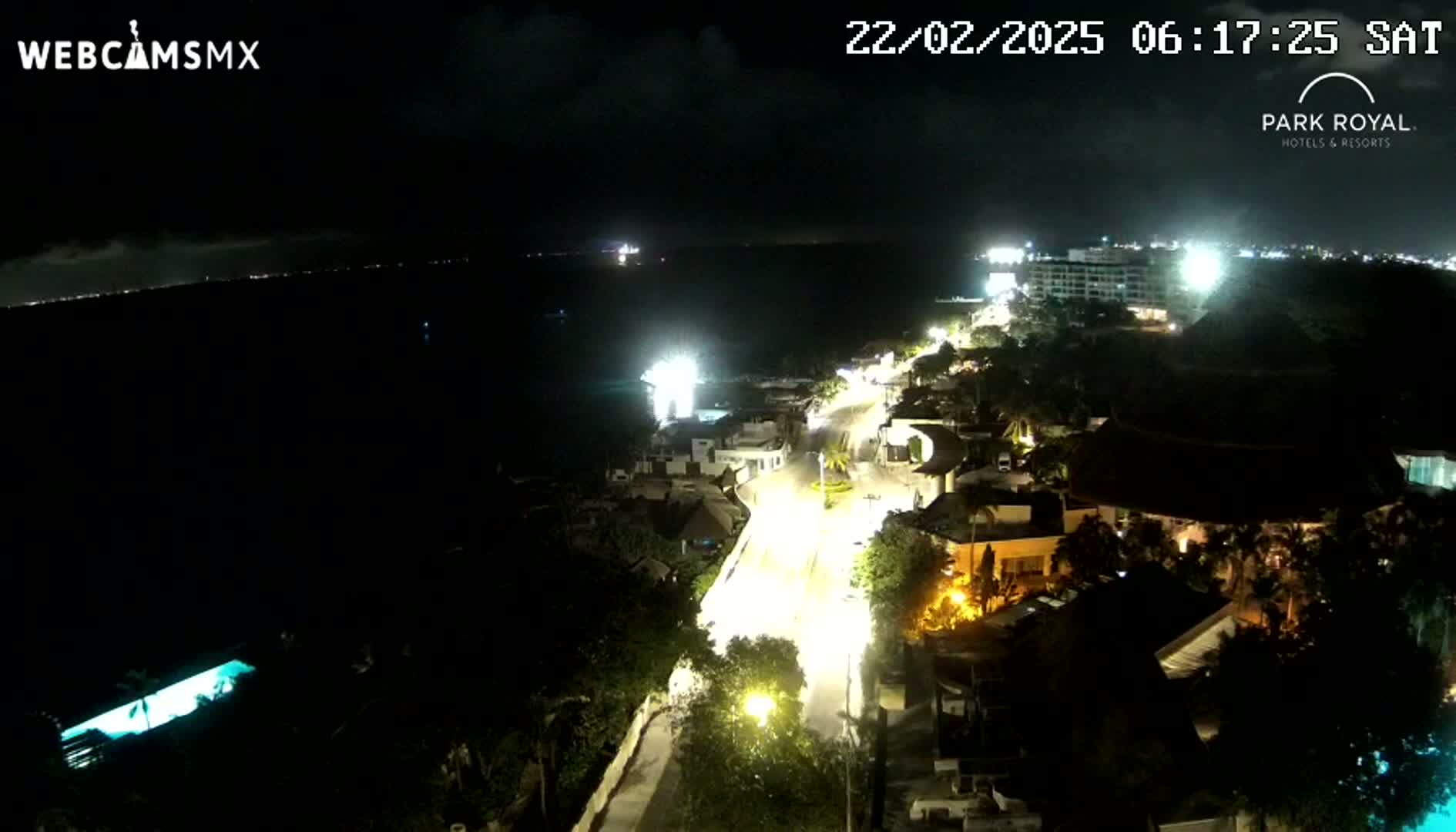 A nighttime aerial view shows a coastal town with brightly lit streets and buildings next to a dark ocean under a partly cloudy sky.