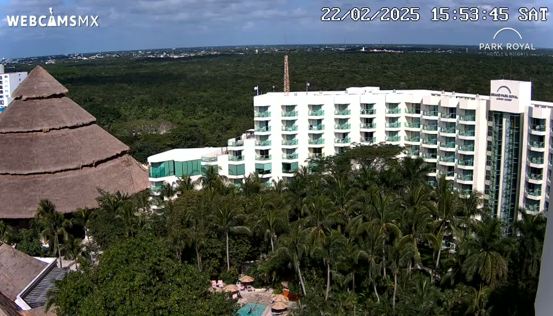 A large hotel complex with a thatched-roof structure sits amidst lush tropical vegetation under partly cloudy skies.