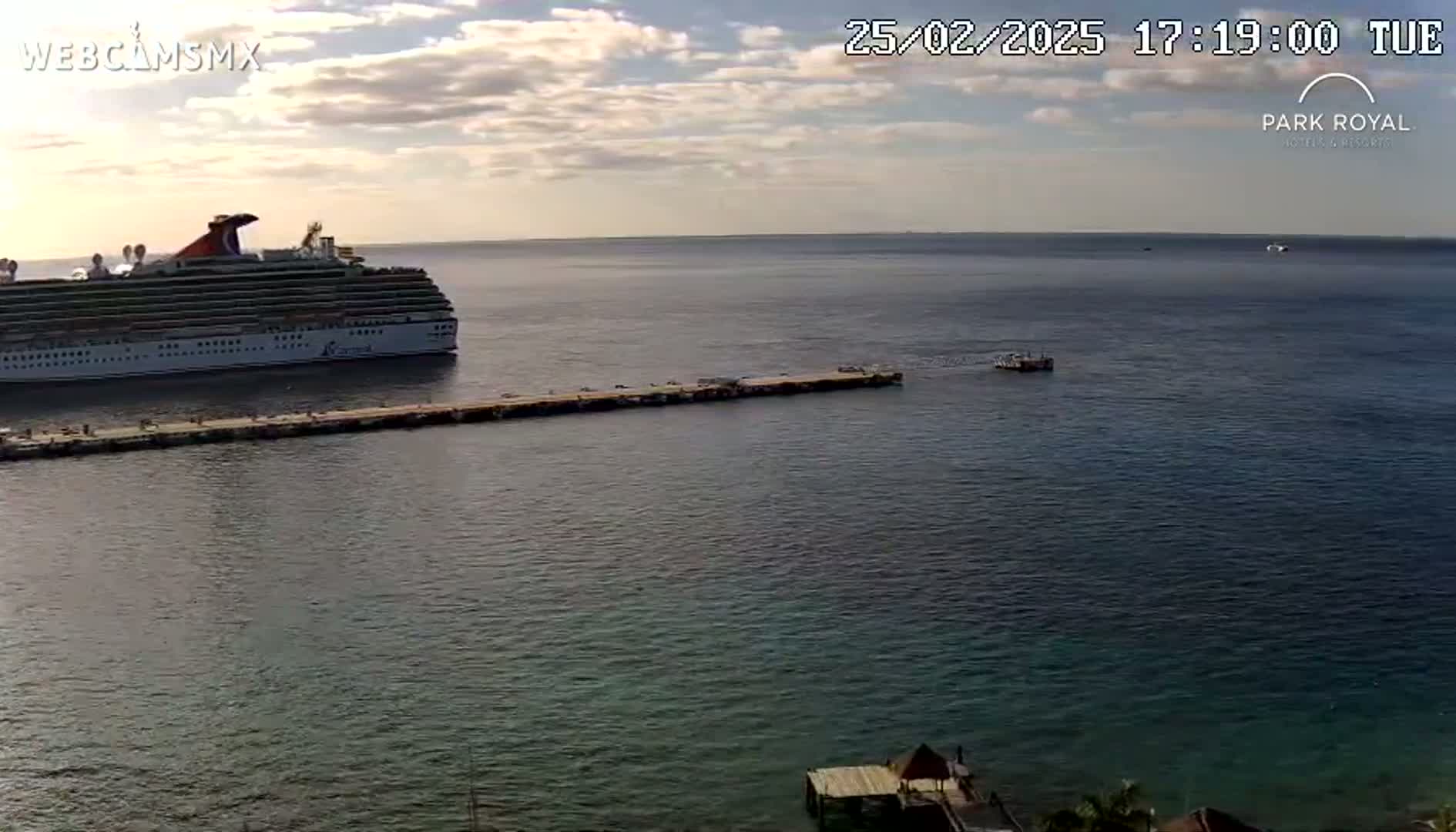 A large cruise ship is docked at a pier on a partly cloudy day, with calm, dark-blue water surrounding it.