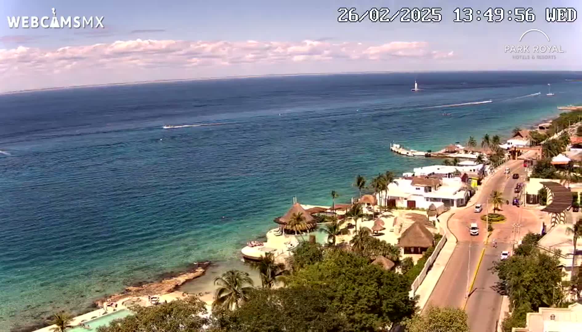 An aerial view shows a coastal town with a road next to a calm, turquoise ocean under a partly cloudy sky.