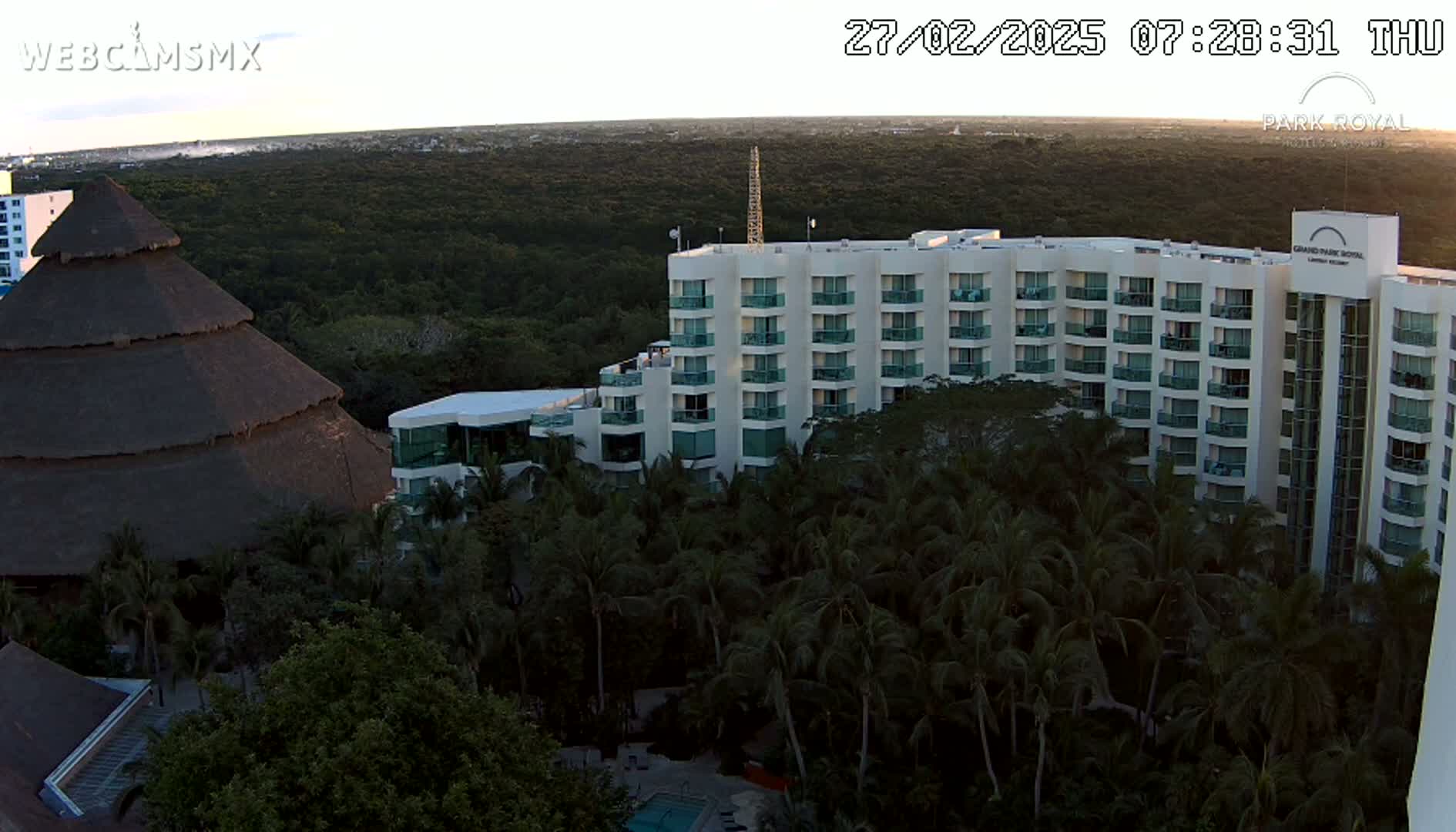 A large, multi-story hotel stands amidst lush tropical foliage at sunrise, with a smaller thatched-roof structure nearby.