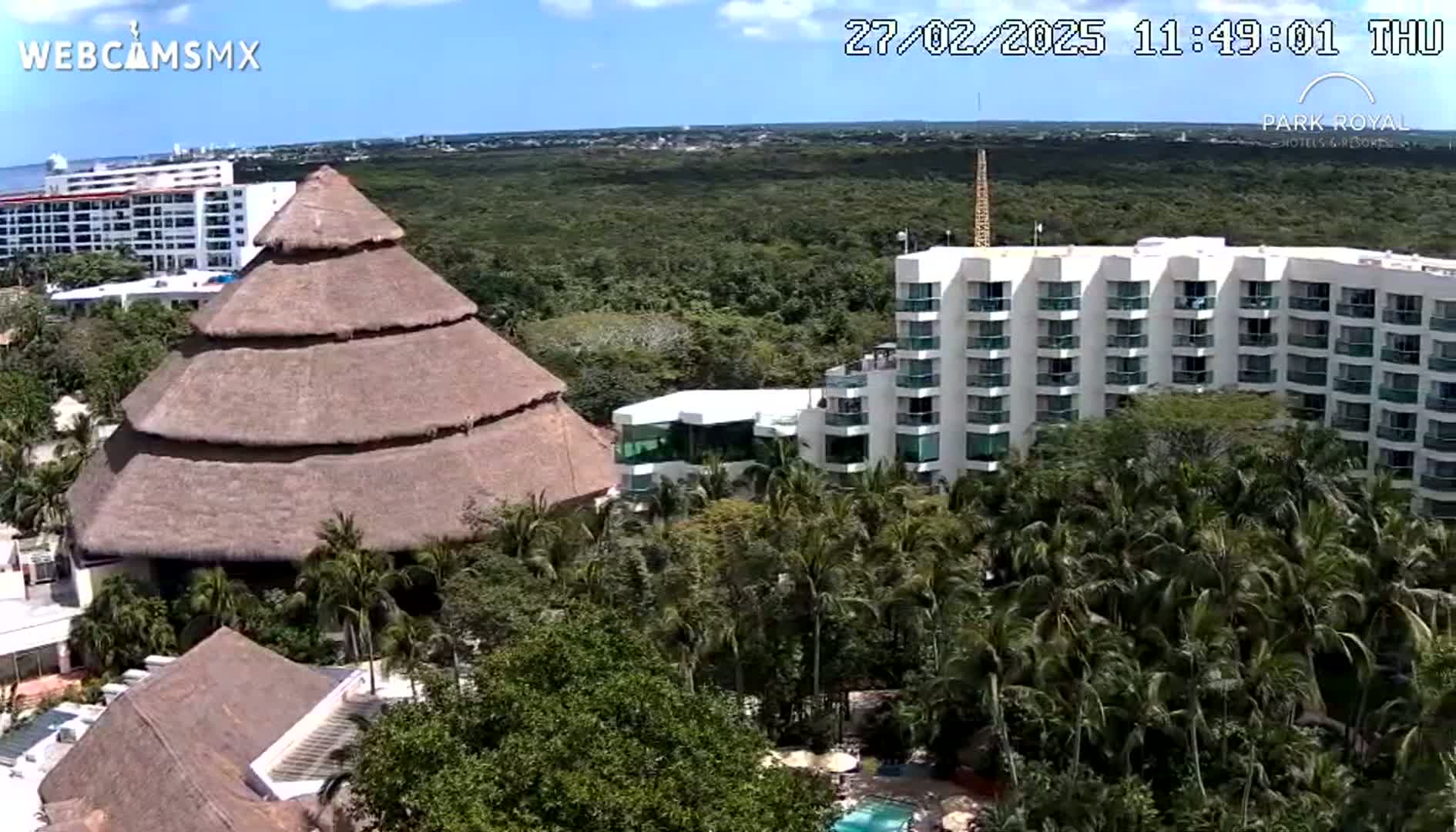 A large, thatched-roof structure sits amidst lush tropical vegetation and a multi-story white hotel building under a mostly sunny sky.