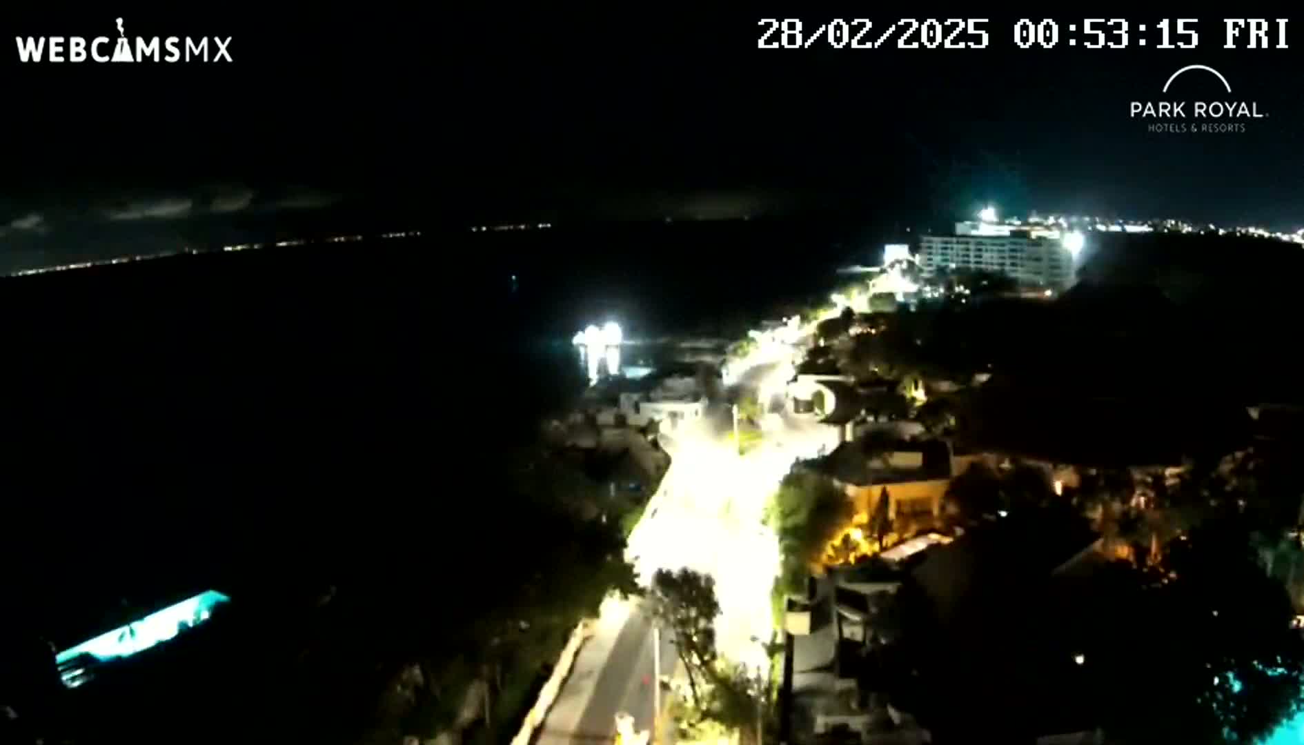 A nighttime, high-angle view shows a coastal road illuminated by streetlights winding through buildings towards a dark body of water with faintly visible lights on the far shore.