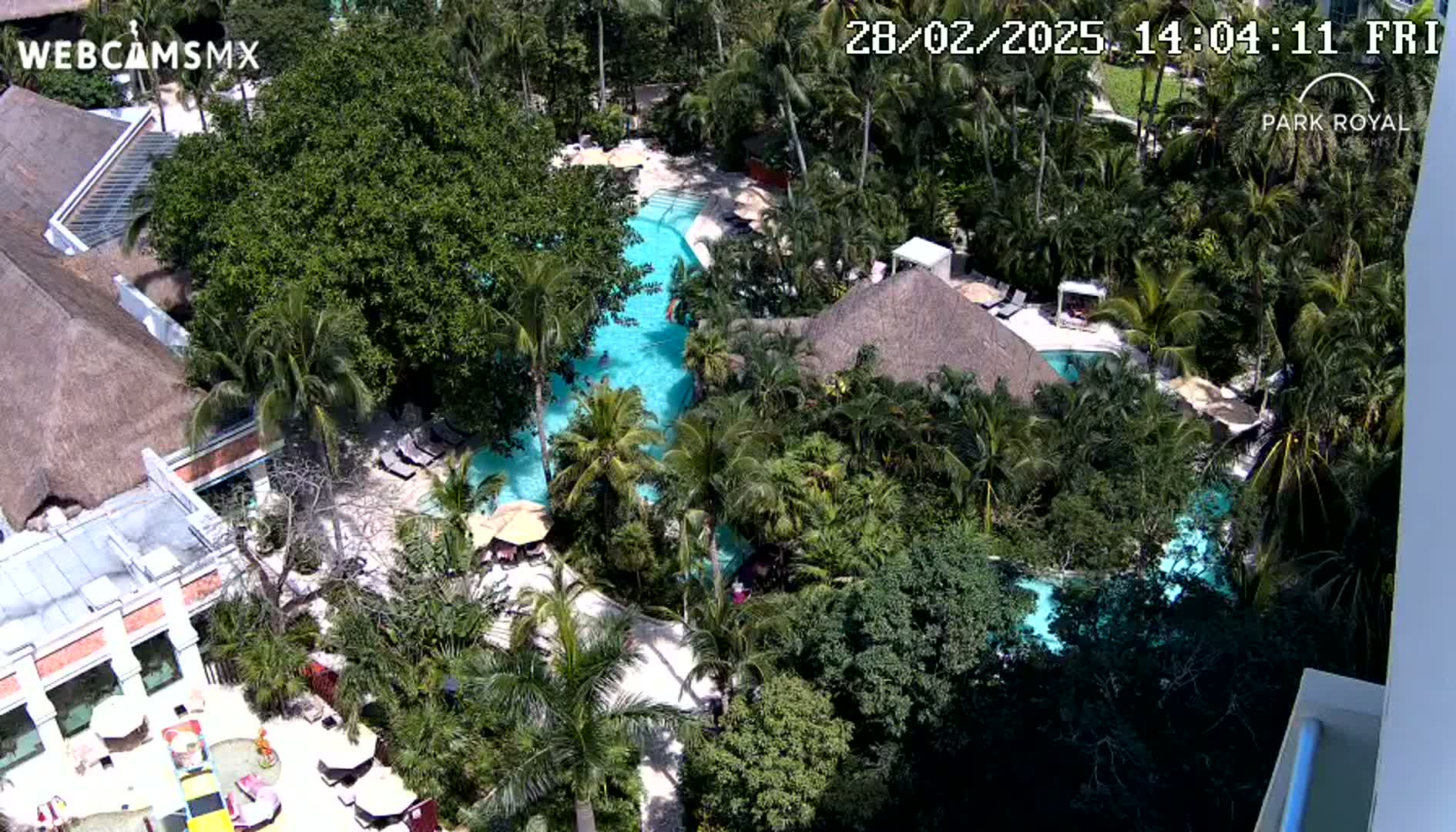 An aerial view shows a resort with multiple pools surrounded by lush tropical vegetation on a sunny day.