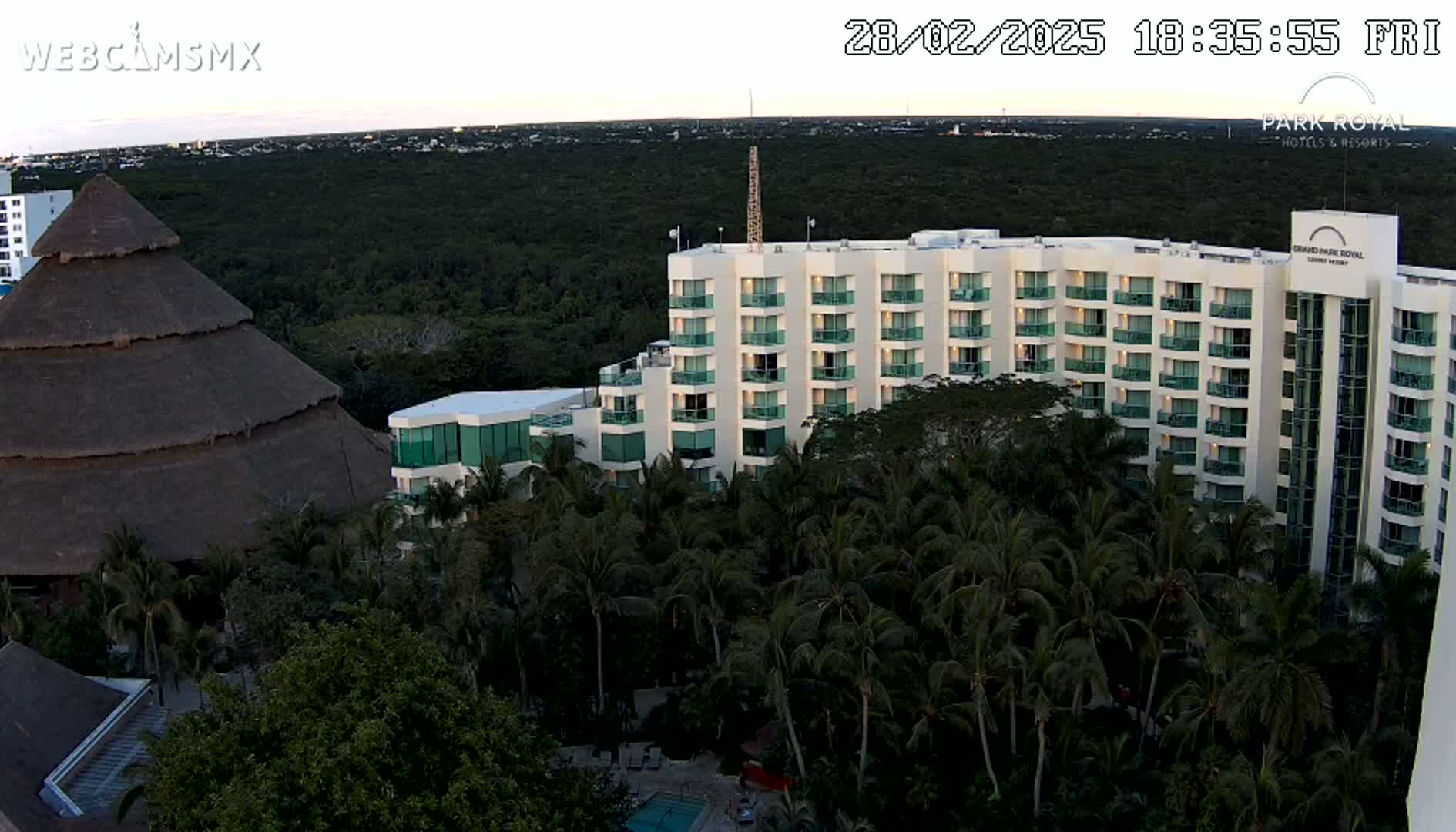 A large hotel complex with numerous palm trees and a thatched-roof structure is nestled within a lush, green landscape under a twilight sky.