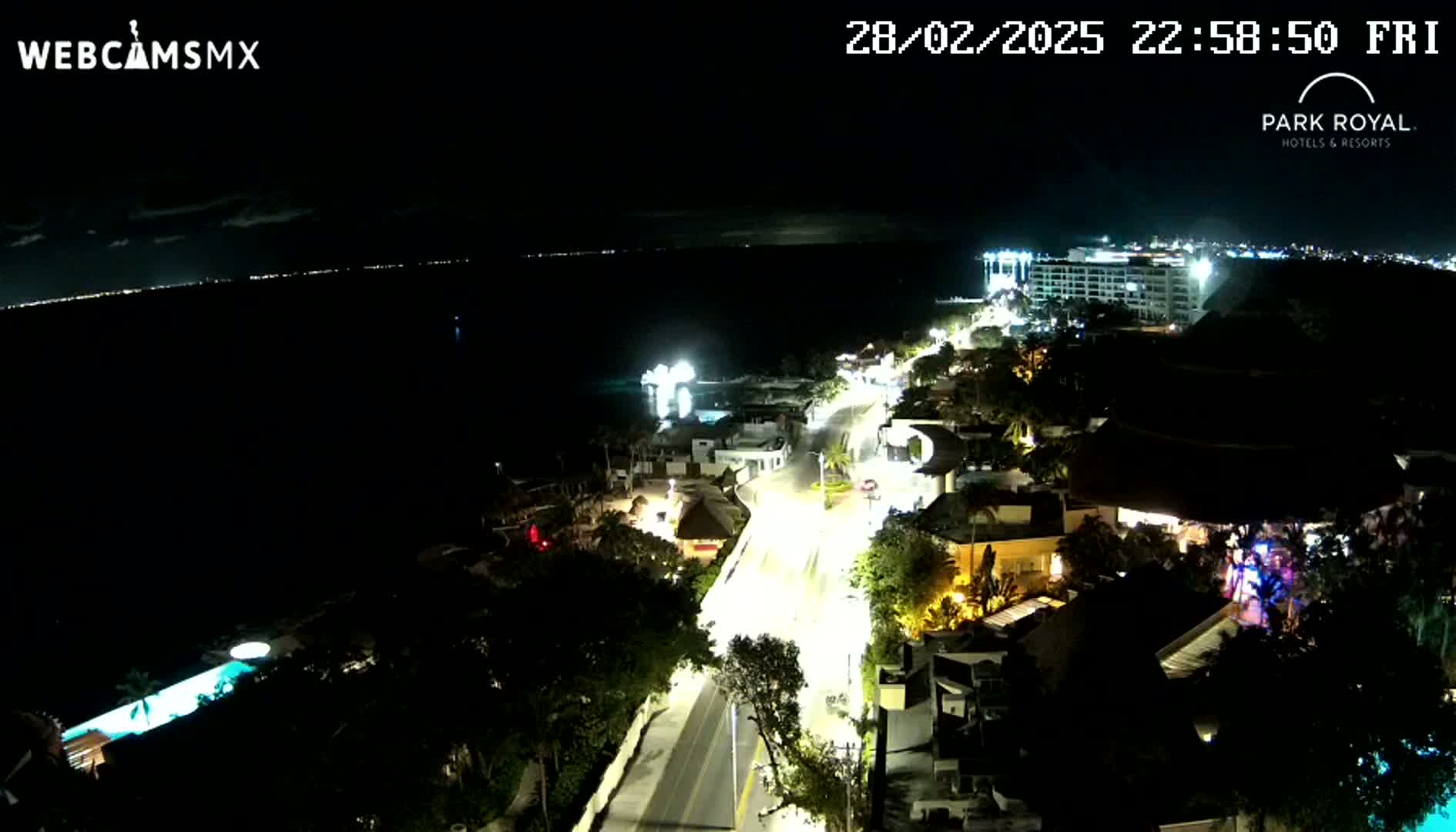 A nighttime aerial view shows a coastal town with illuminated streets and buildings next to a dark body of water under a clear sky.
