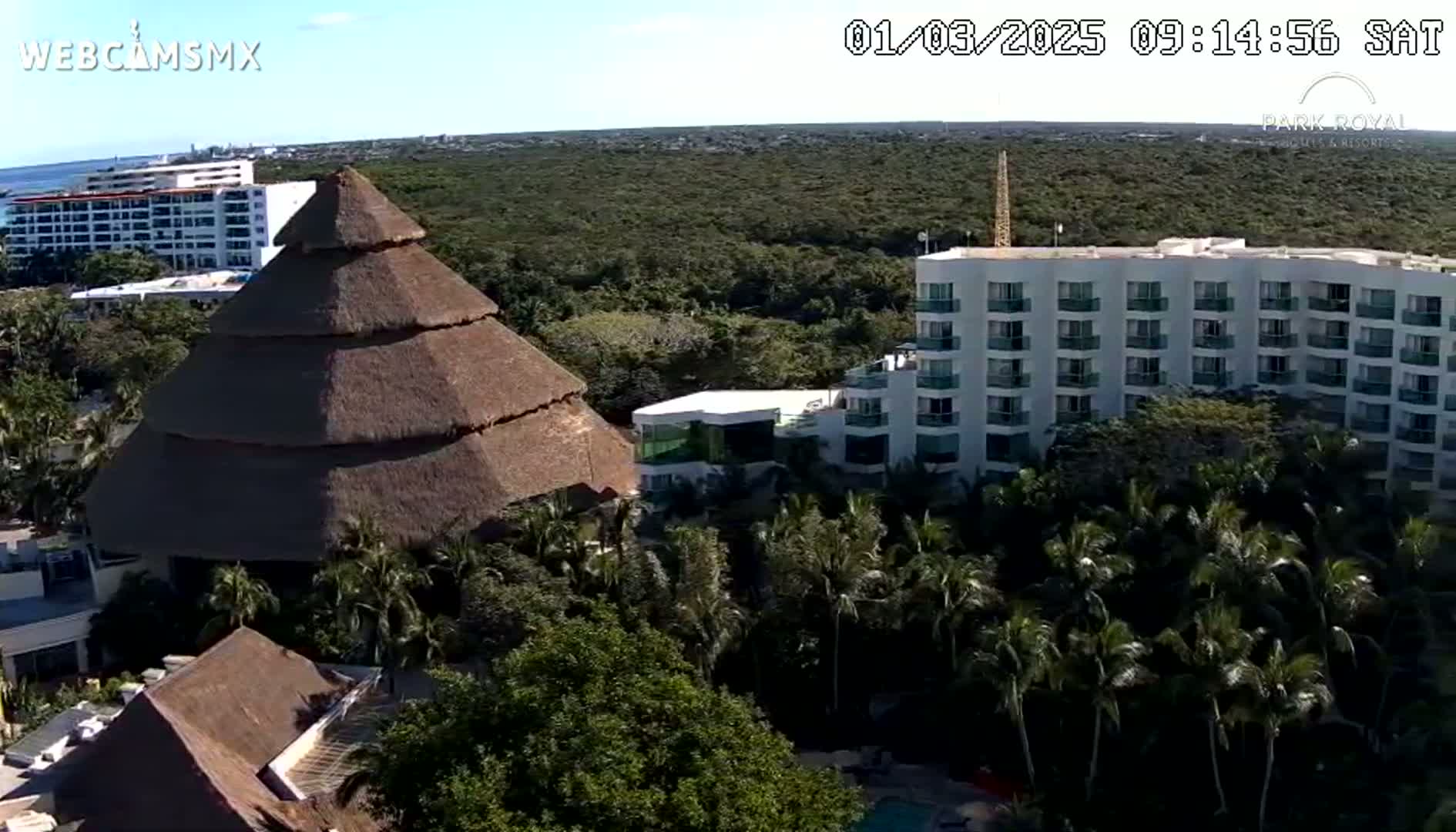 An aerial view shows a resort with thatched-roof buildings and a multi-story hotel nestled amongst lush green vegetation under a clear, sunny sky.