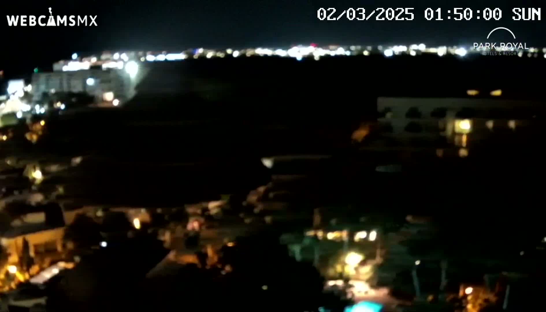 A nighttime, blurry, high-angle view of a city illuminated by lights, with a distant cityscape visible in the background.