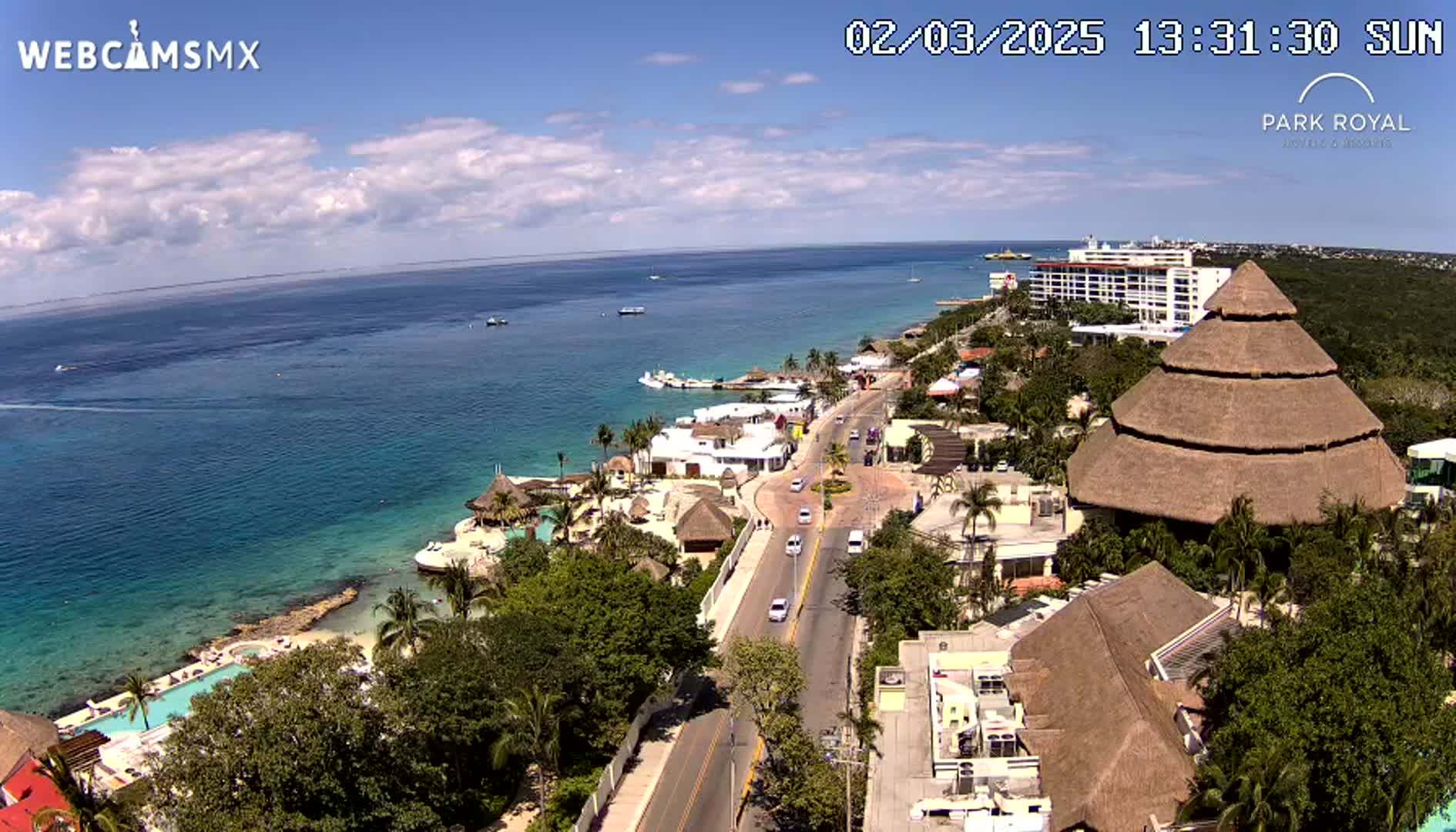 An aerial view of a coastal resort town on a sunny day shows a road leading from the ocean to buildings, including a large thatched-roof structure.