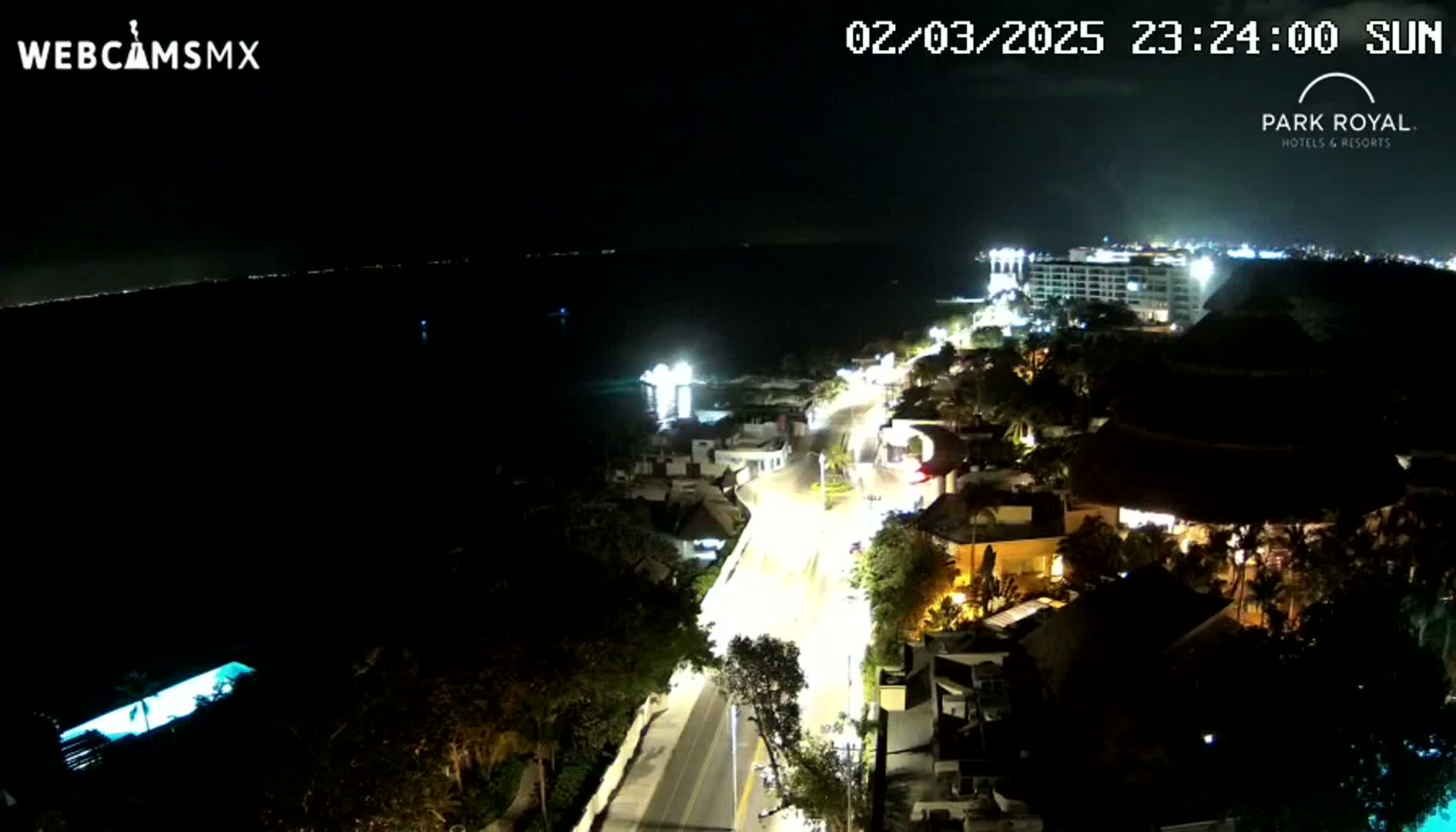 A nighttime, aerial view shows a coastal road with illuminated buildings and vehicles, leading to a large resort near a dark body of water under a clear sky.