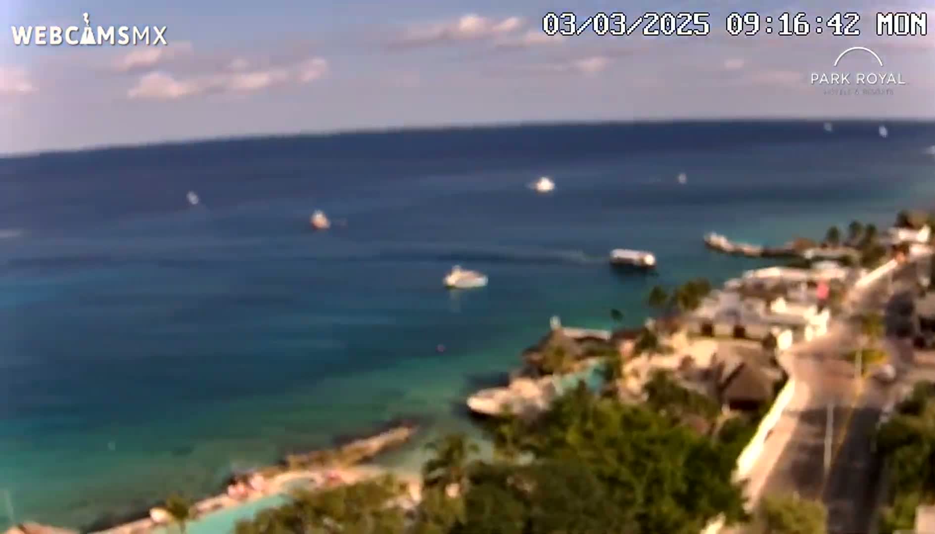 An elevated view of a tropical coastline on a partly cloudy day shows a calm ocean with several boats, a resort with lush vegetation, and a road winding through the property.