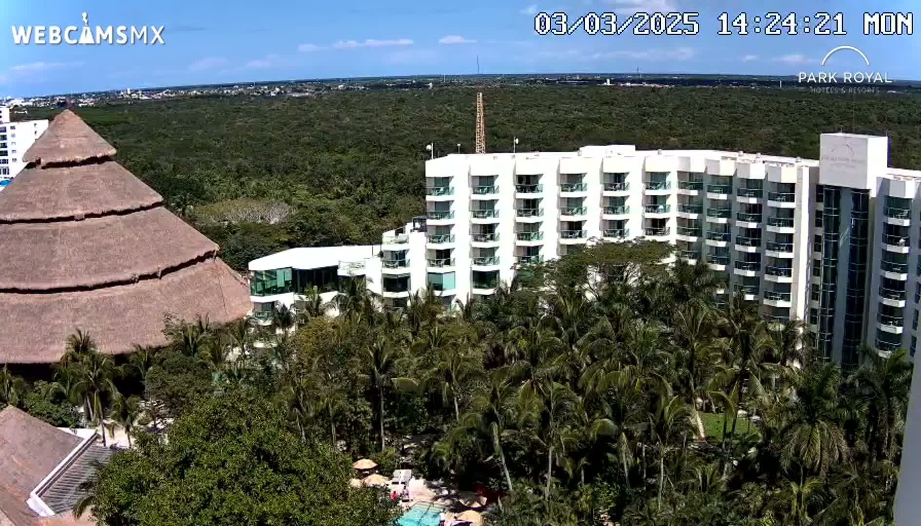 A large resort complex with a thatched-roof building and a multi-story hotel sits amidst lush green tropical vegetation under a mostly sunny sky.
