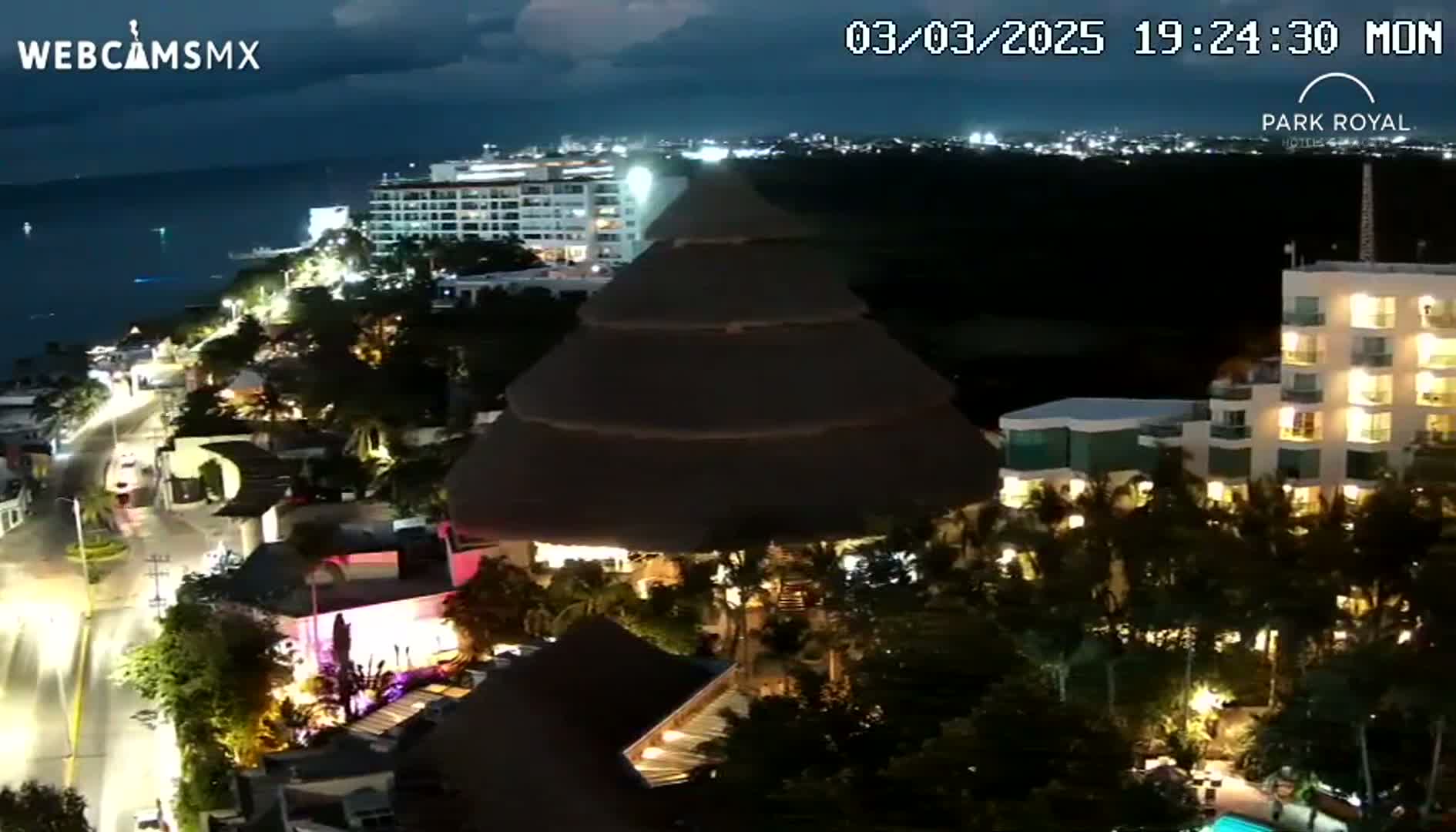 A nighttime aerial view shows a coastal town with illuminated buildings, palm trees, and a dark ocean under a partly cloudy sky.