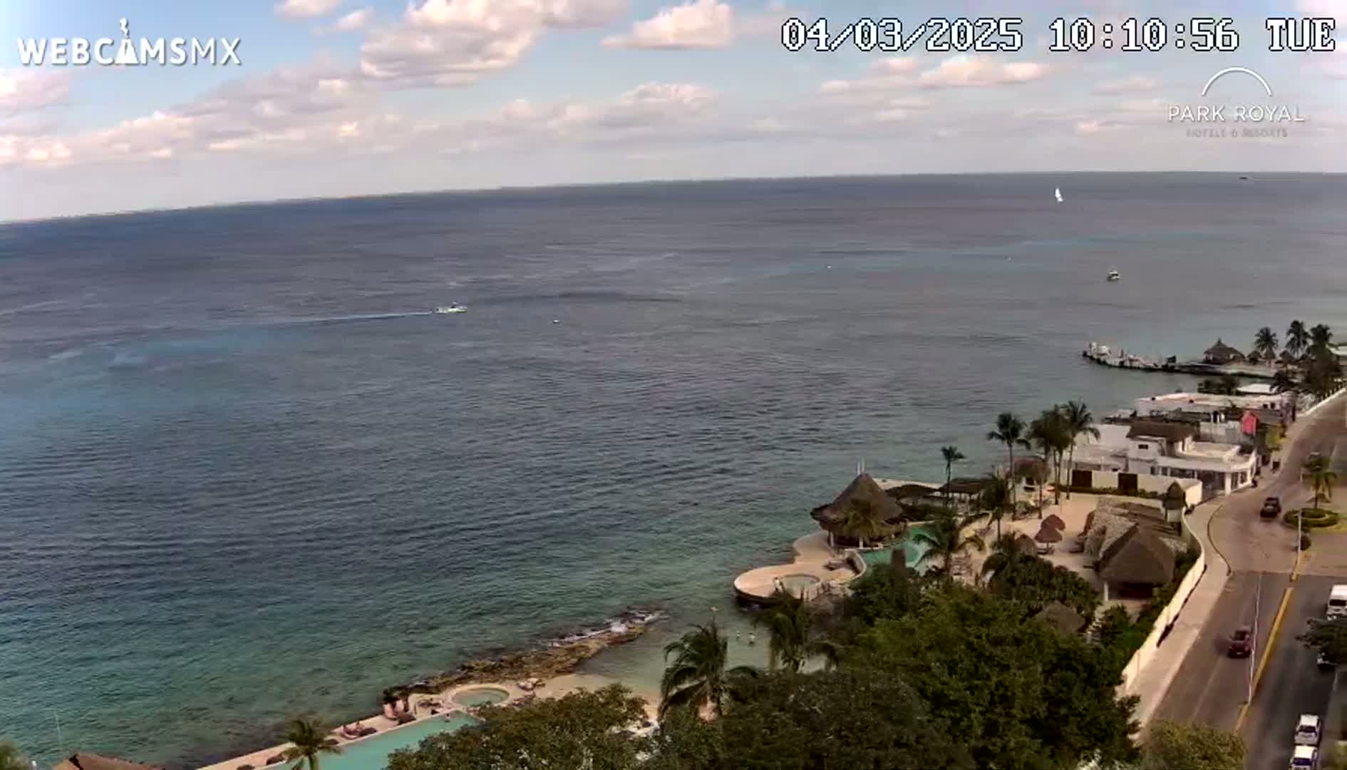 An expansive ocean view on a partly cloudy day shows a resort with palm trees, thatched roof structures, a swimming pool, and a road with cars next to the water.