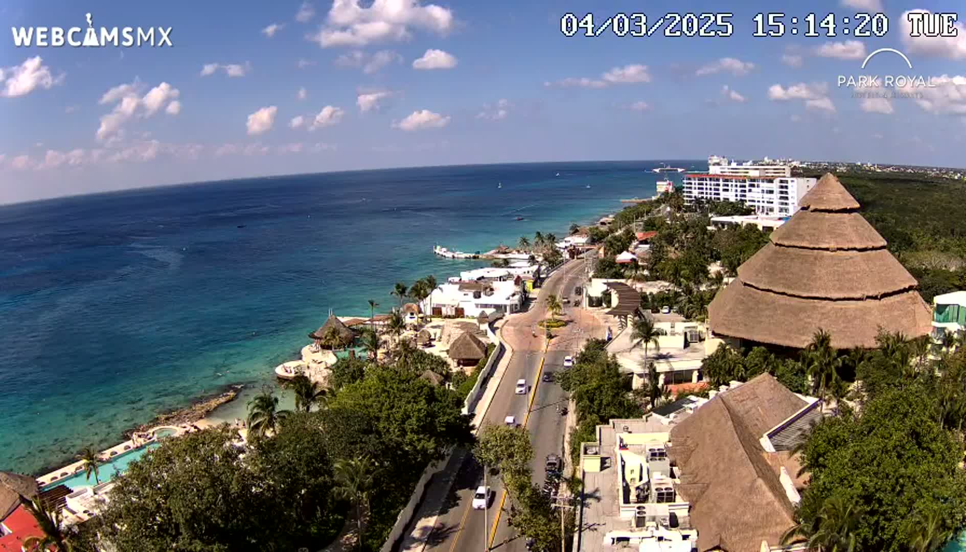 An aerial view shows a coastal resort town on a sunny day with a clear blue ocean and a road winding through the buildings toward the water.