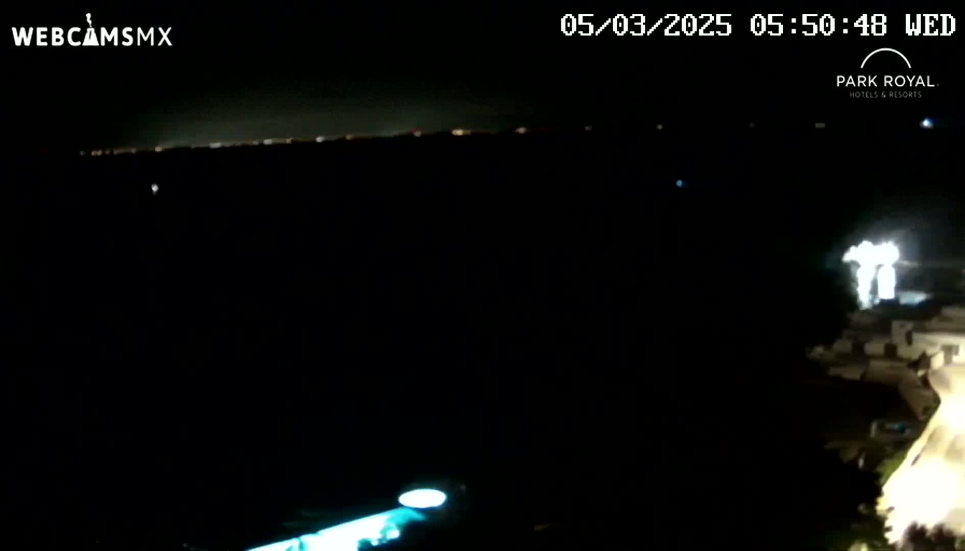 A nighttime long shot shows a distant city skyline illuminated against a dark sky, with a brightly lit structure and swimming pool visible in the lower right corner.