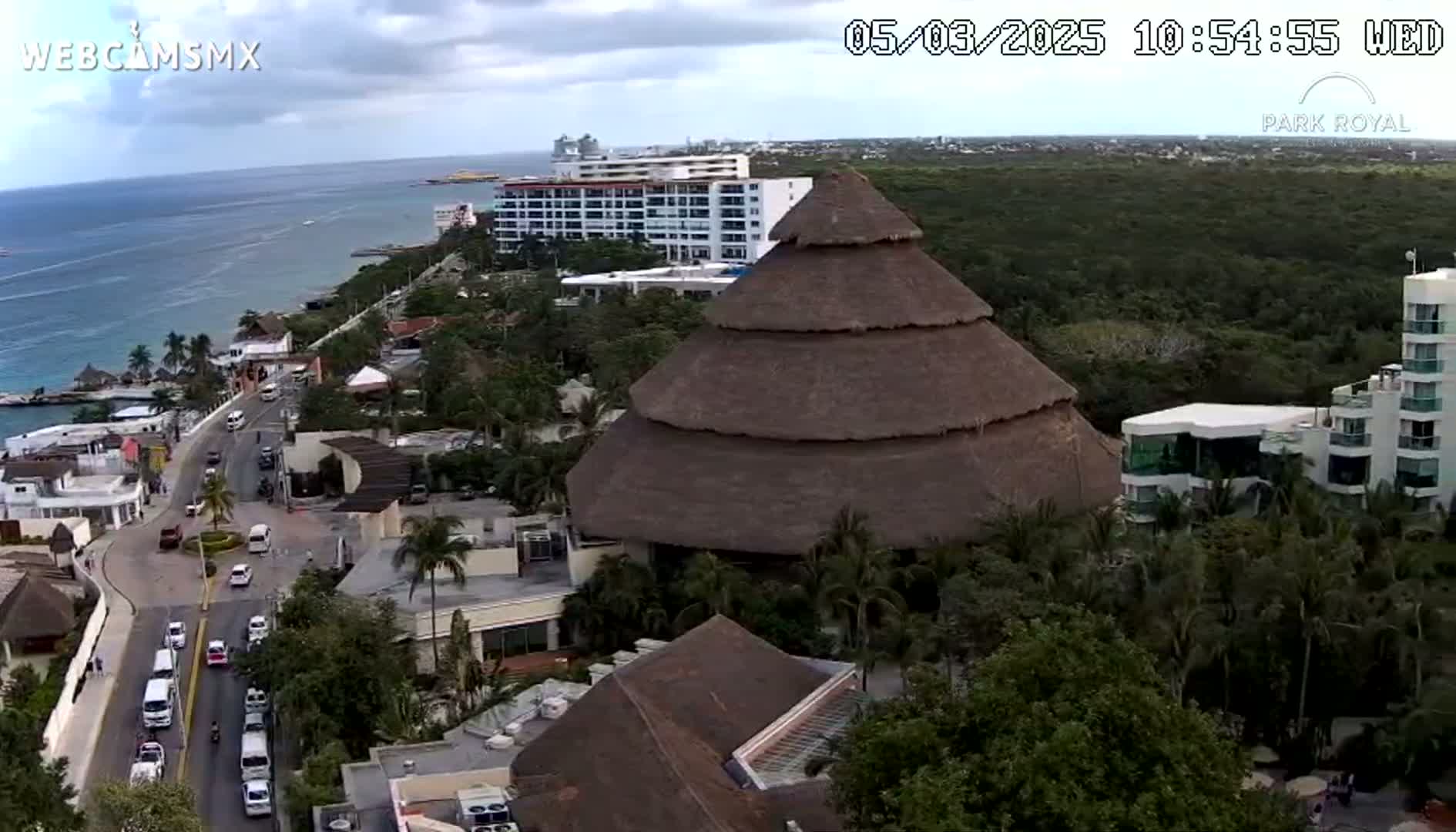 An aerial view shows a large thatched-roof structure near a coastline with overcast skies, overlooking a road with cars and multiple buildings, including hotels.