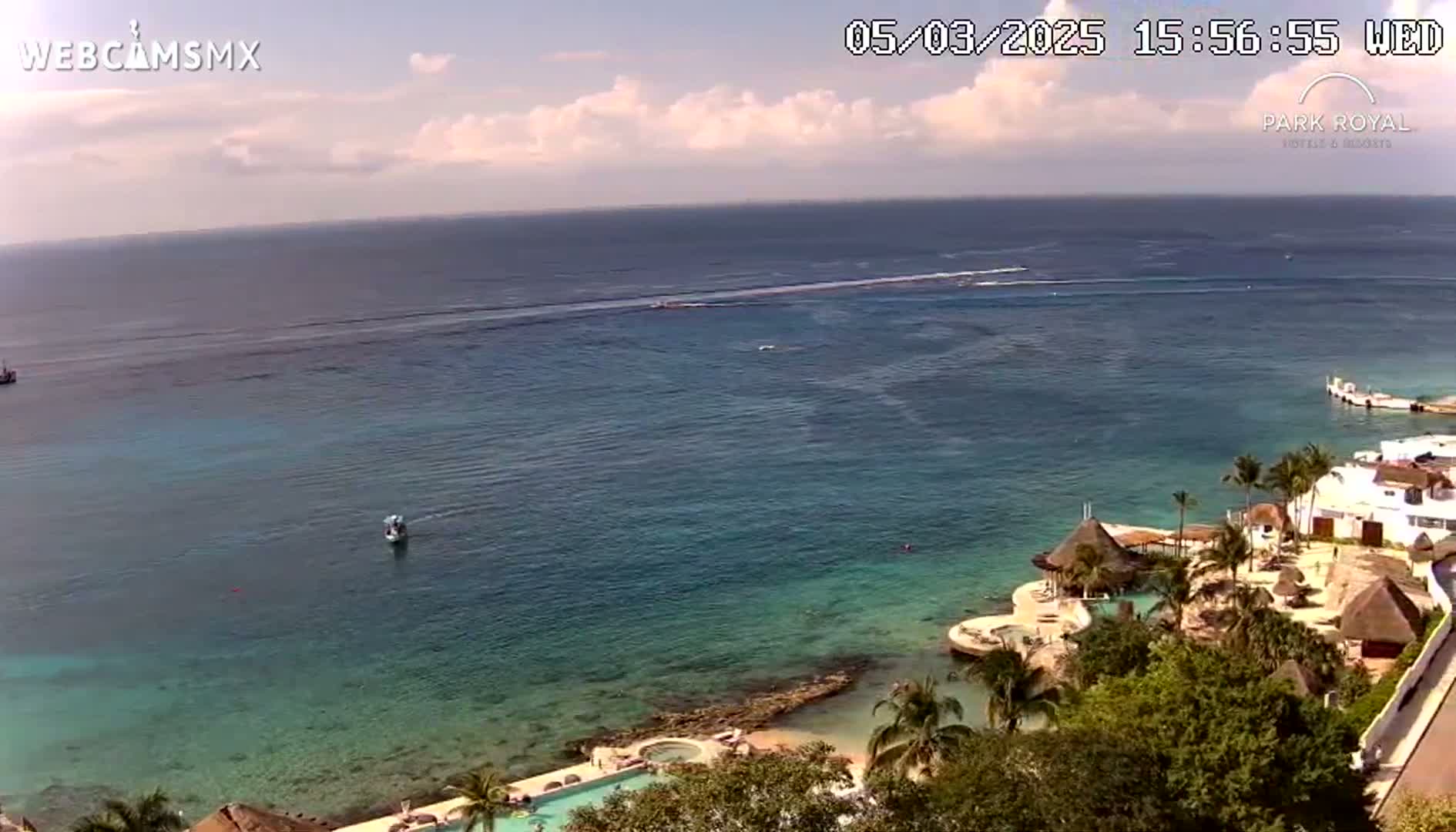 A partly cloudy day shows a resort on a coast with clear turquoise water, a small boat in the distance, and palm trees surrounding the buildings.
