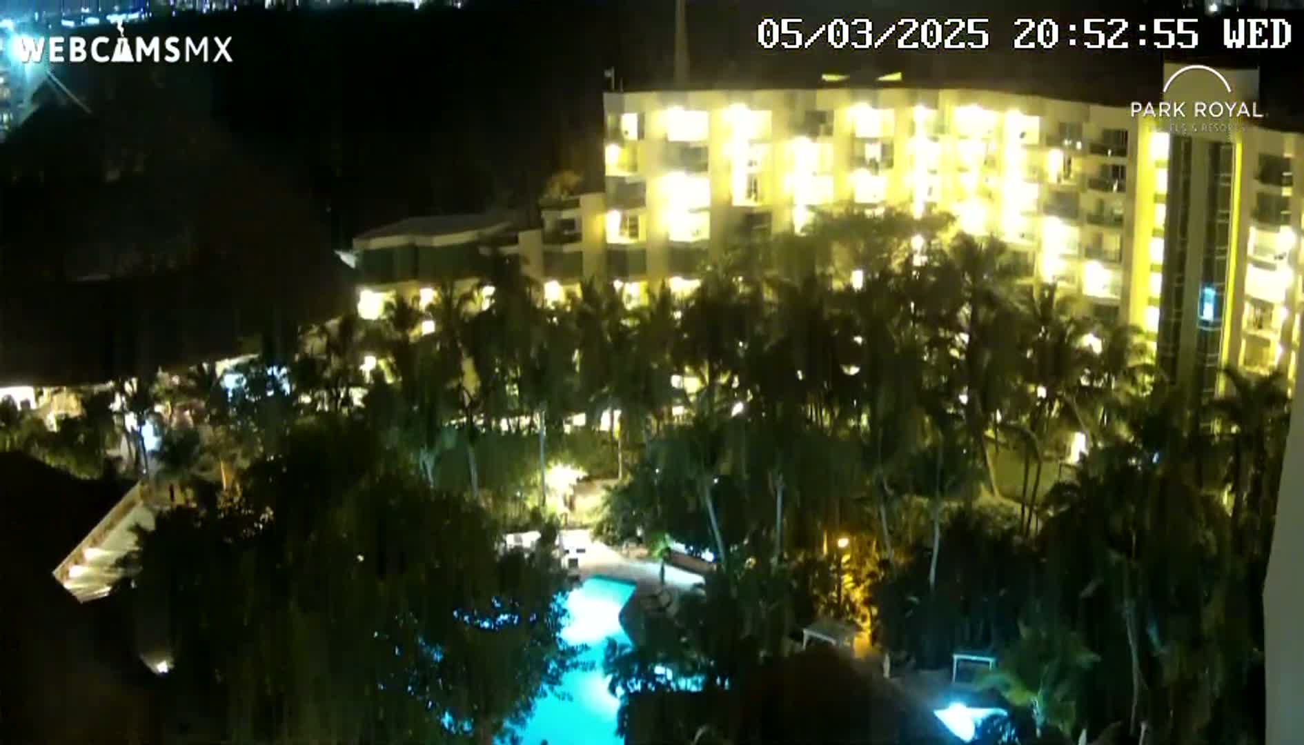 A brightly lit hotel at night, surrounded by palm trees and a swimming pool, under a dark sky.