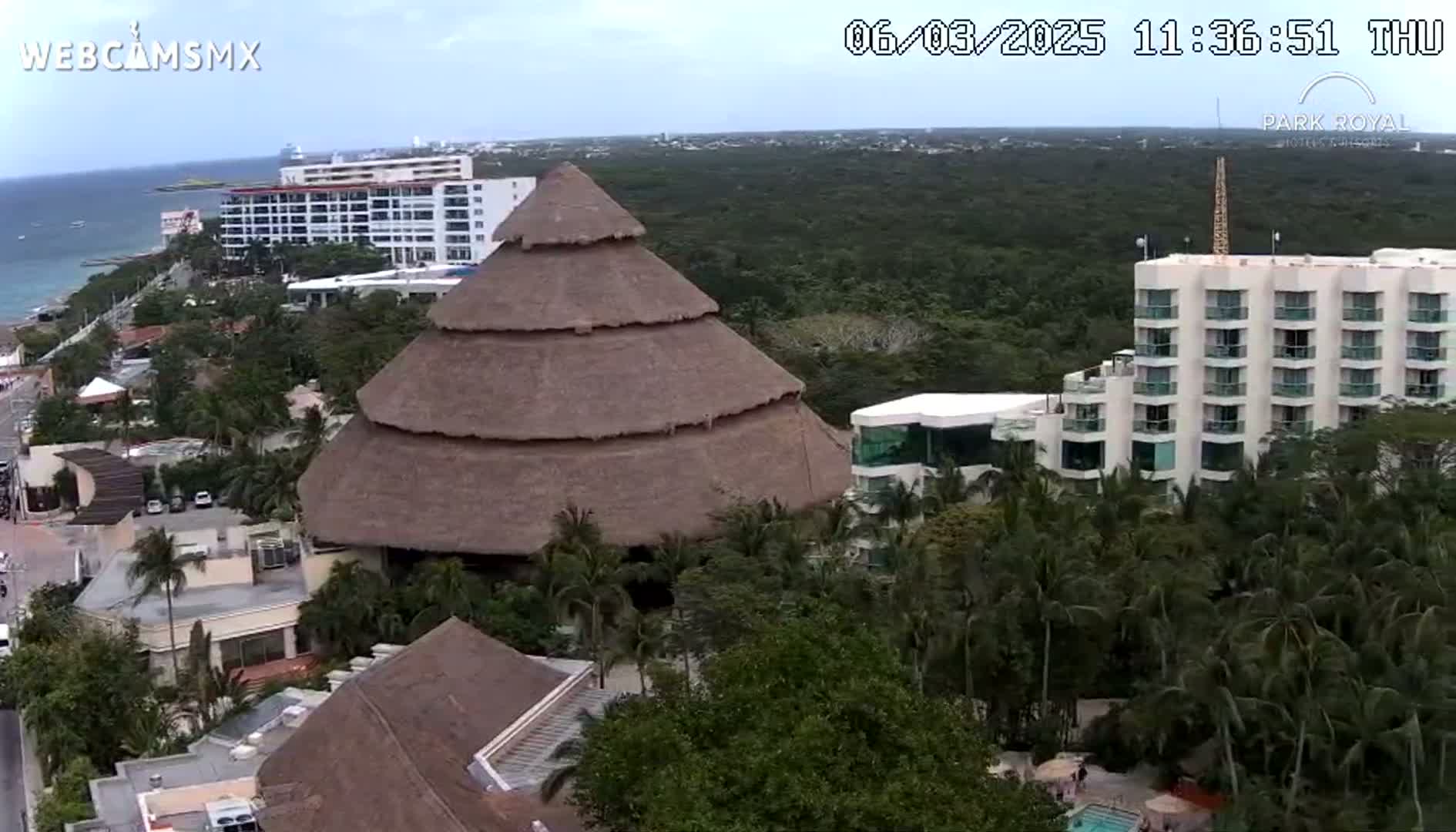 An aerial view shows a large thatched-roof structure and several multi-story buildings surrounded by lush tropical vegetation near an ocean under partly cloudy skies.
